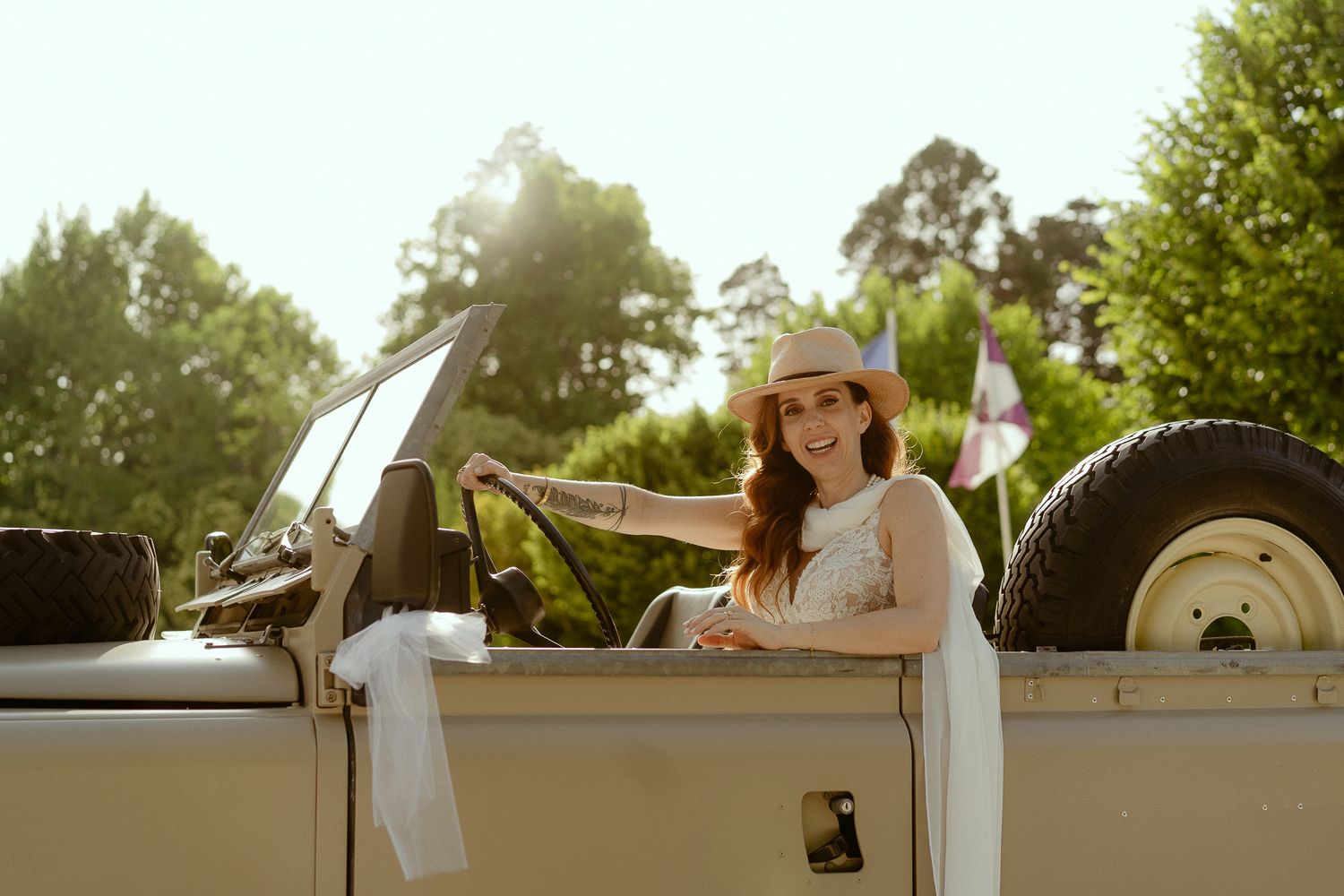 Smiling woman in a straw hat sitting in a vintage open-top jeep surrounded by lush green trees.