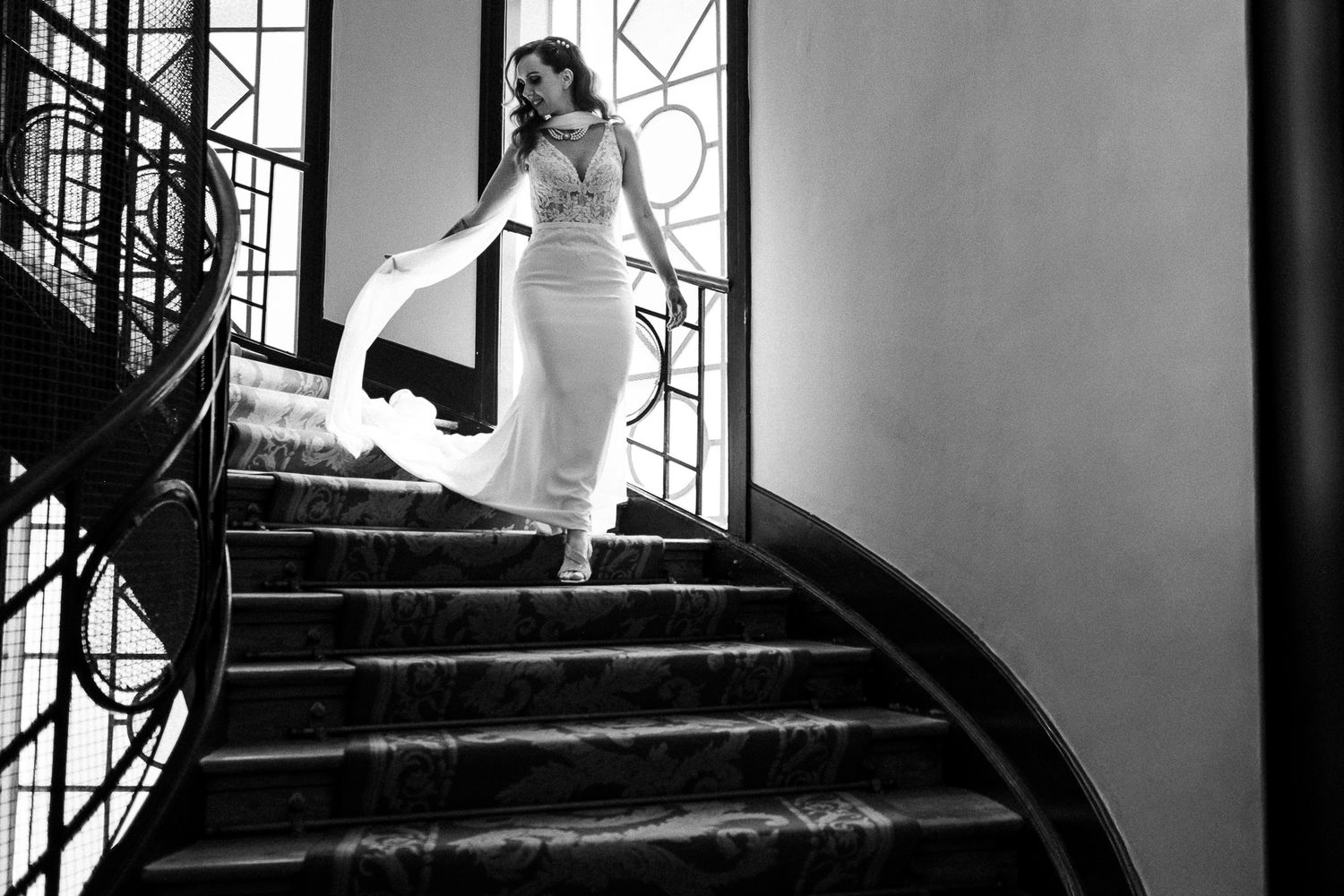 Bride in white gown descending elegant curved staircase in black and white wedding photo.