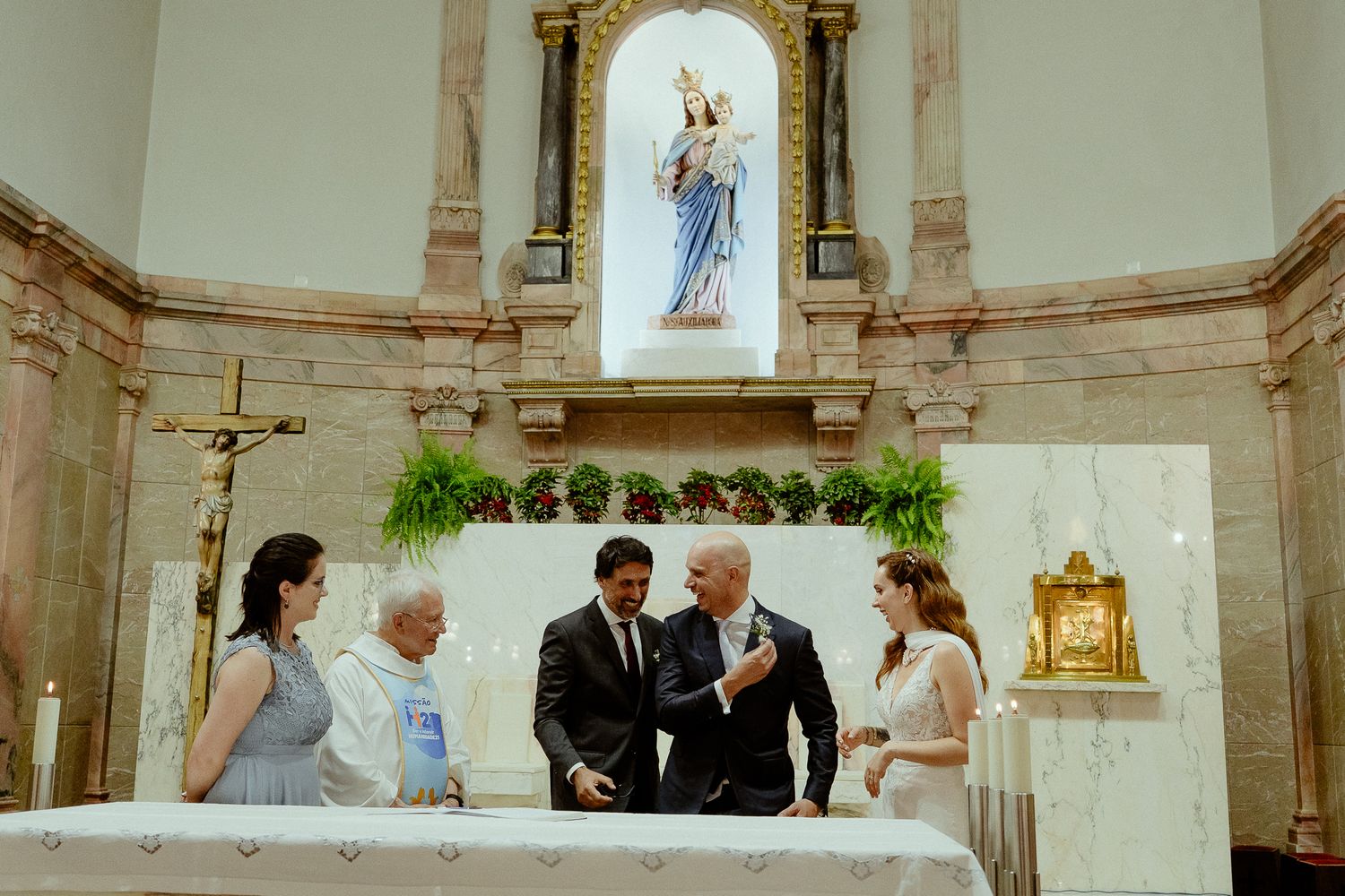Wedding ceremony at a Catholic church altar with bride, groom, and guests gathered before a Virgin Mary statue.