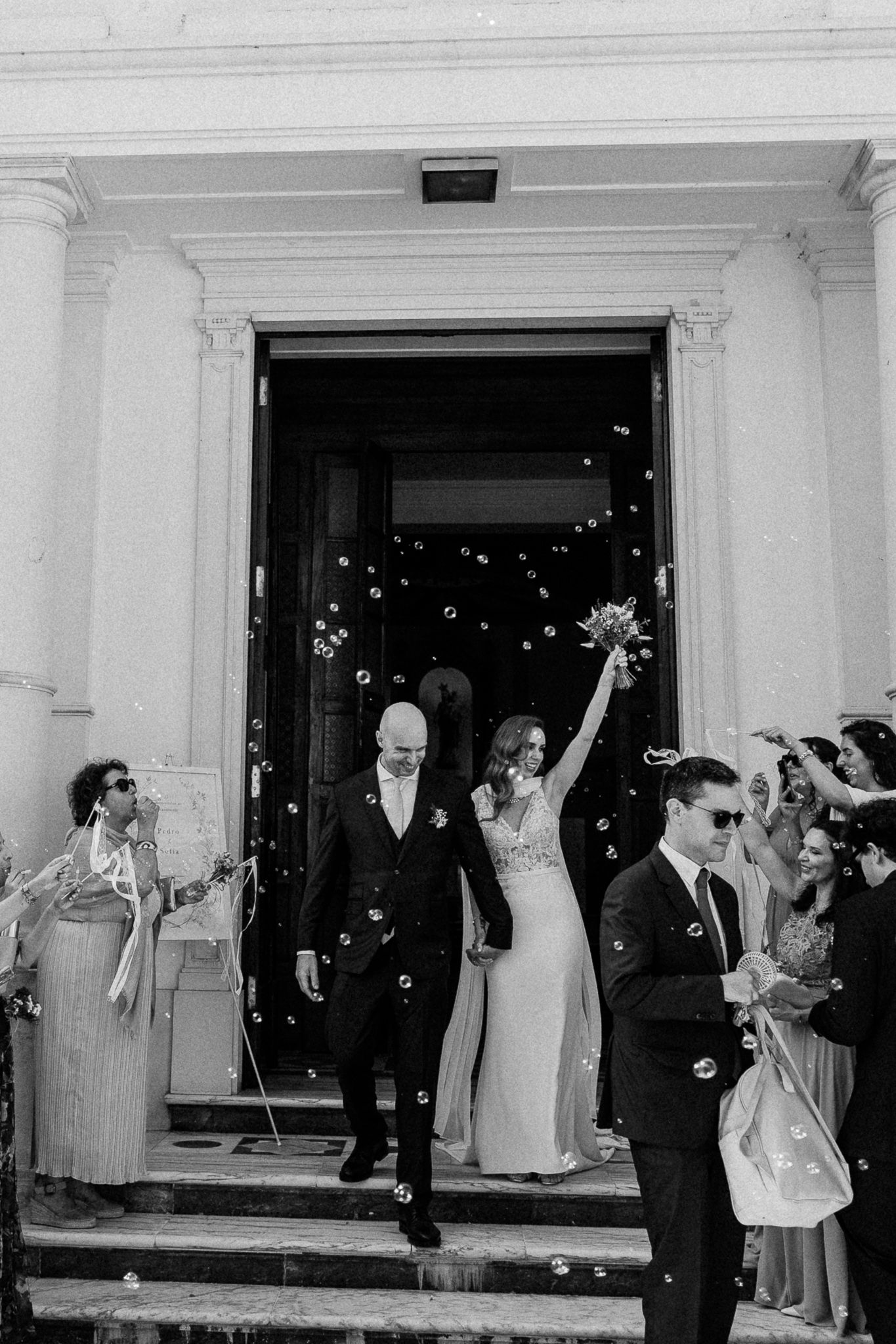 Black and white photo of a bride and groom exiting a church surrounded by guests blowing bubbles.