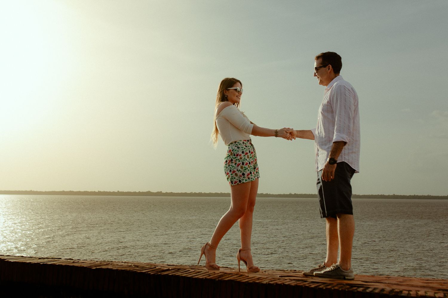 A couple stands together on a wooden pier at sunset, holding hands against a serene ocean backdrop.