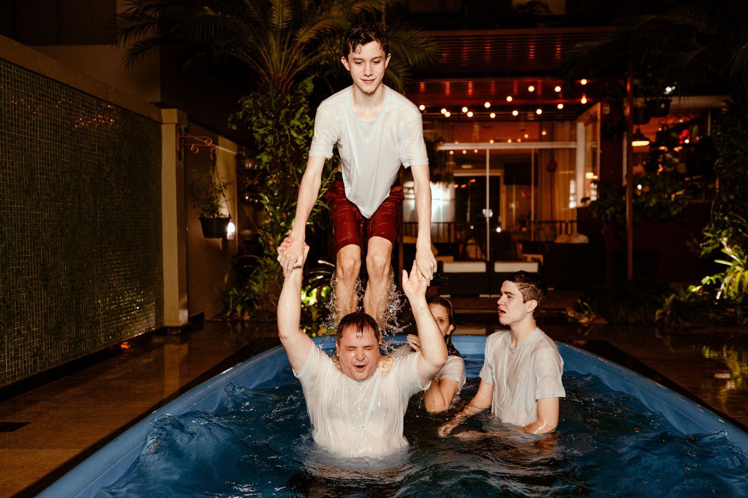 Three people playing and splashing in a pool at night with red string lights illuminating the outdoor space.