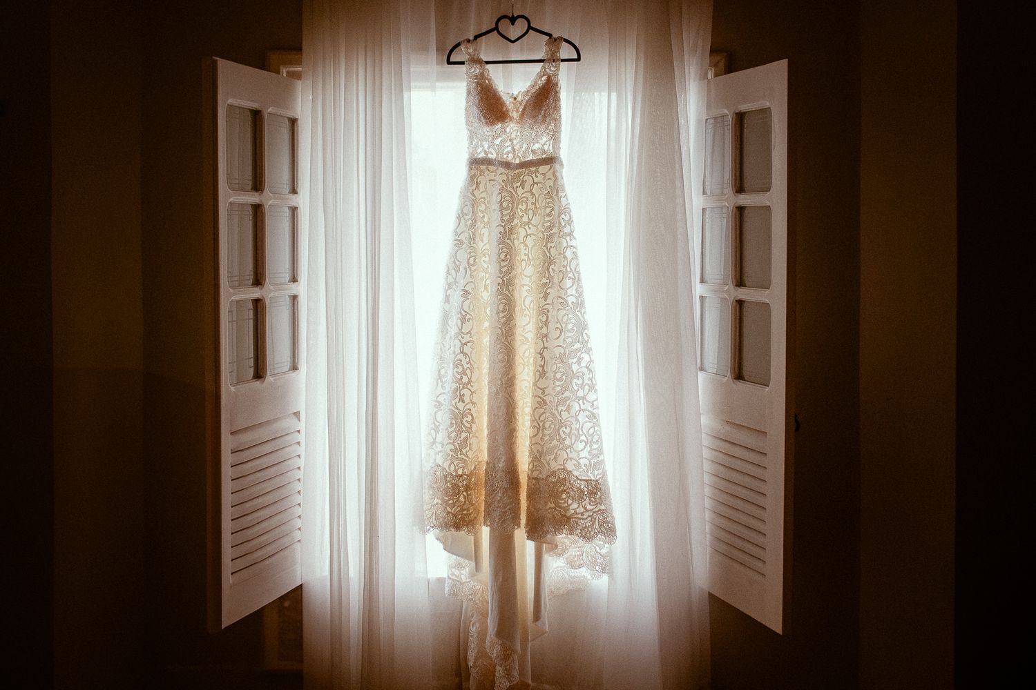 A wedding dress hangs in a window with natural light streaming through sheer curtains, creating a romantic silhouette.