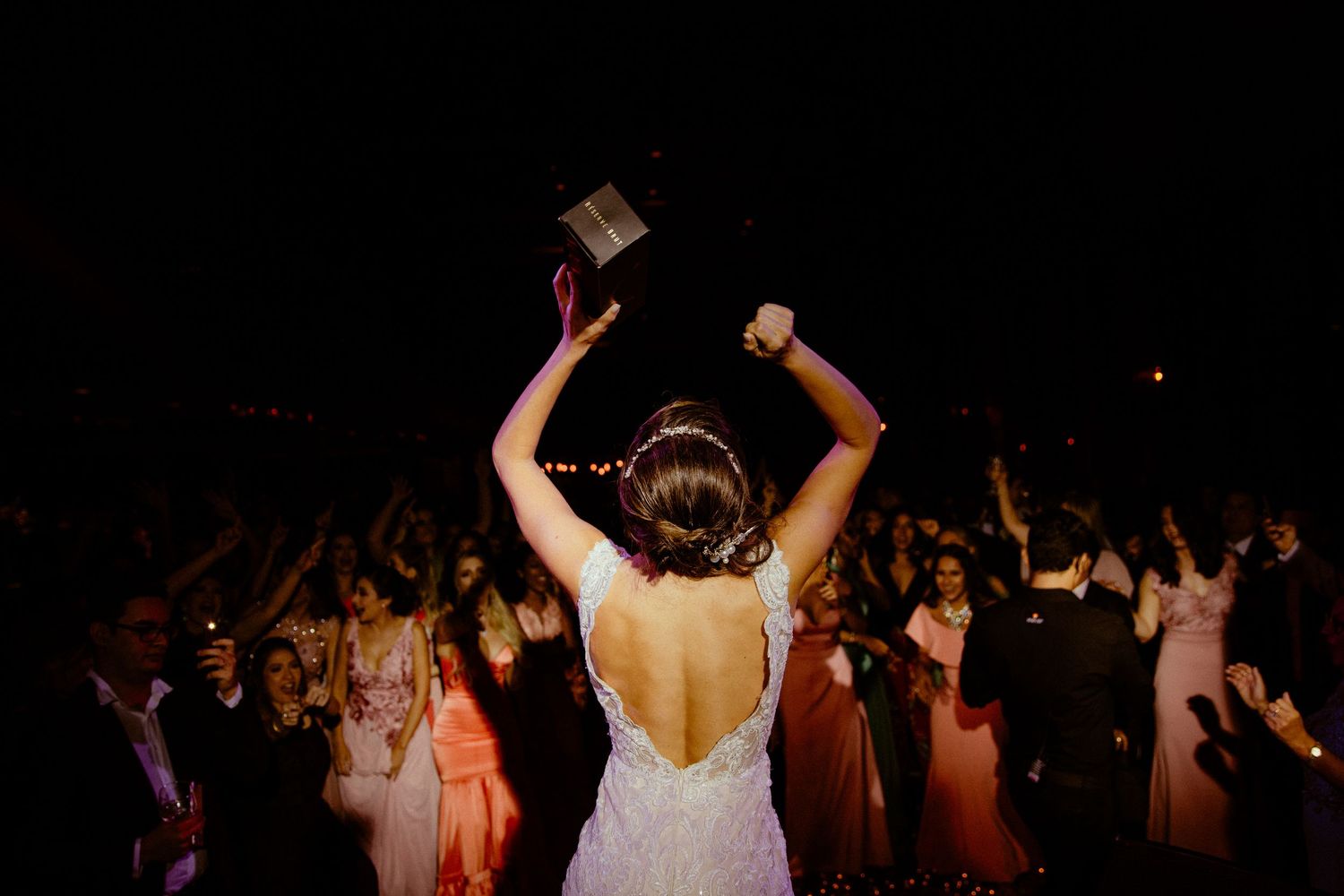 A crowded dance floor at a wedding reception with people dancing and celebrating in a dark venue with ambient lighting.