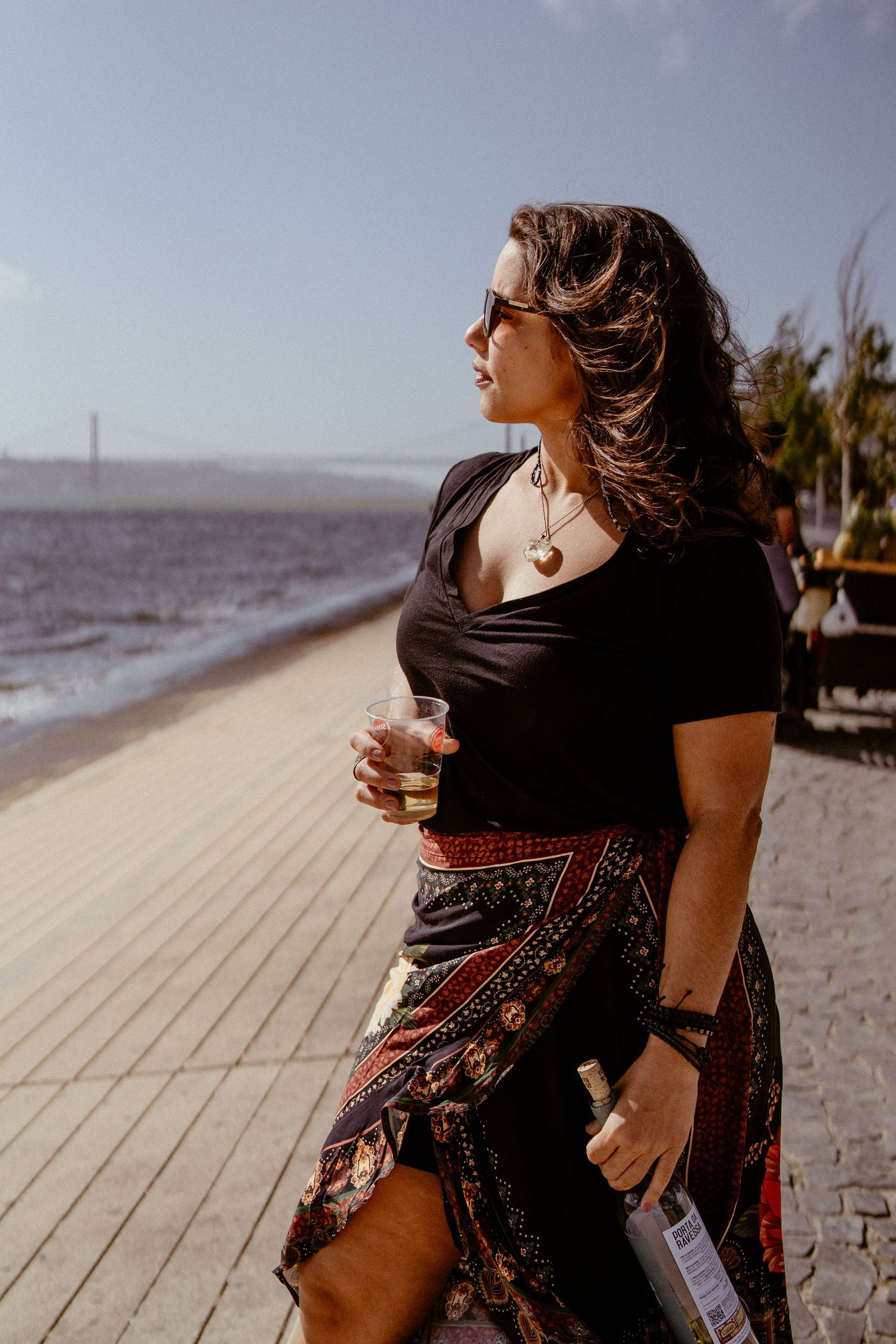 A person wearing a patterned skirt and black top stands on a wooden boardwalk by the ocean on a sunny day.