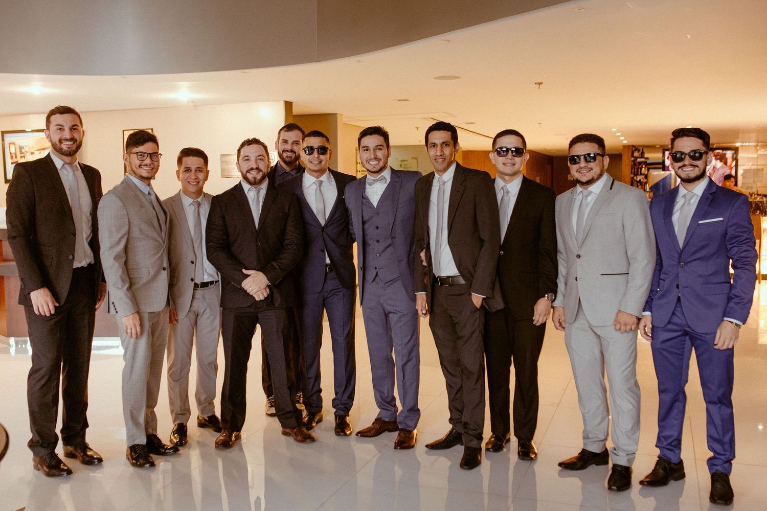 A group of men in formal suits stand together in a line against an indoor wall.
