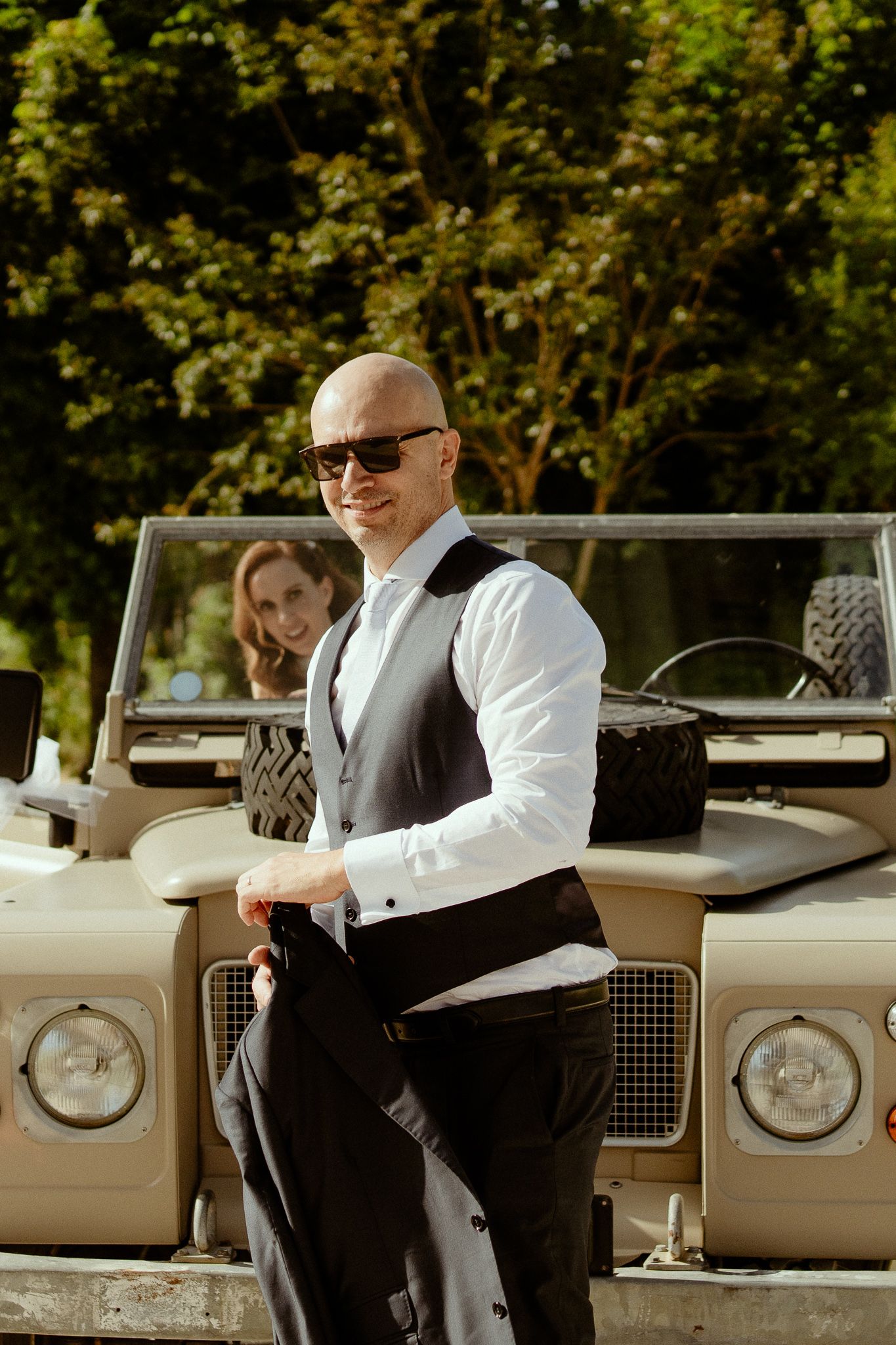 Bald groom in white shirt and gray vest with sunglasses stands by vintage Land Rover on sunny wedding day.