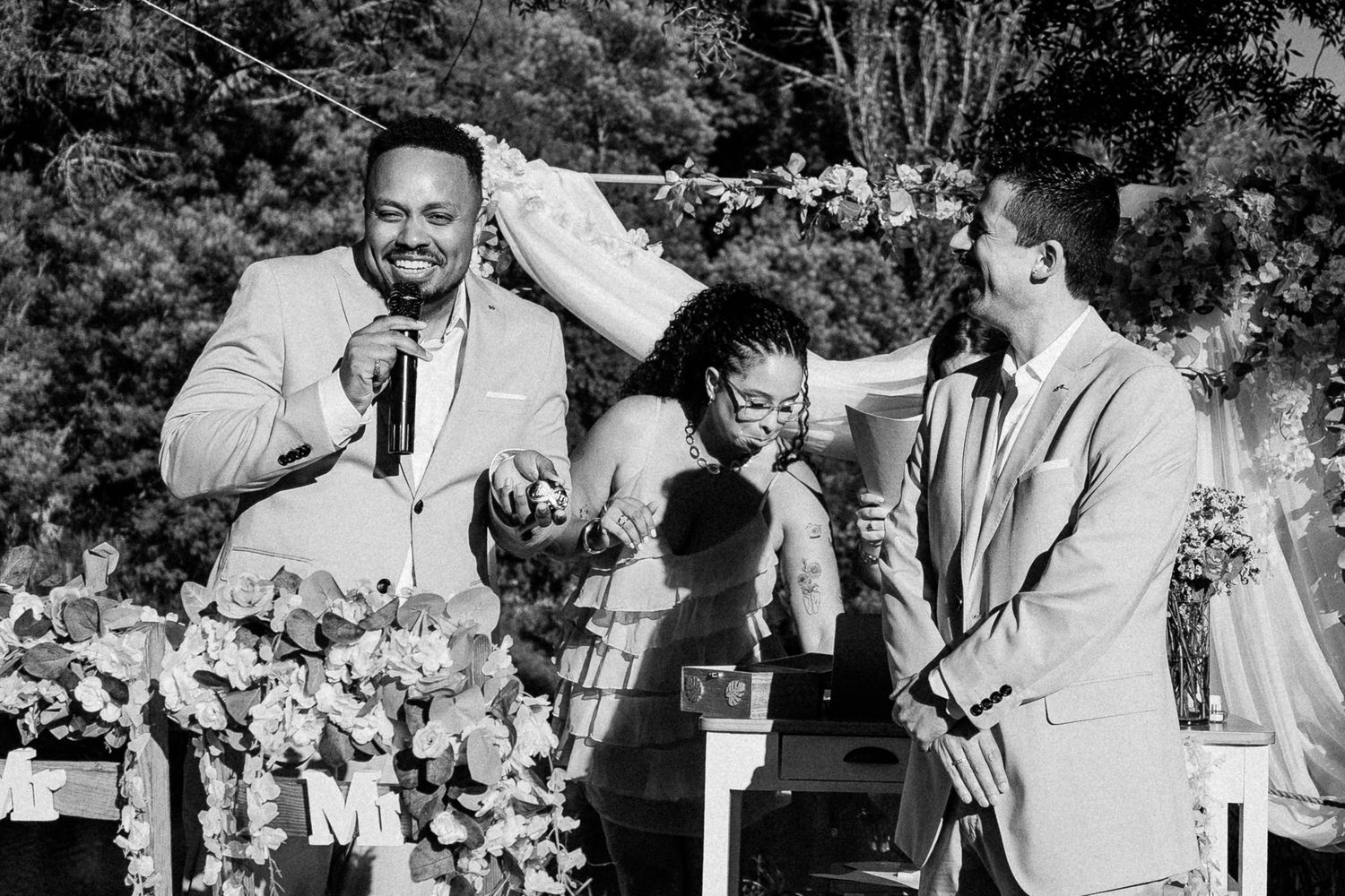 Black and white wedding ceremony photo showing officiant speaking while bride laughs and groom smiles outdoors.