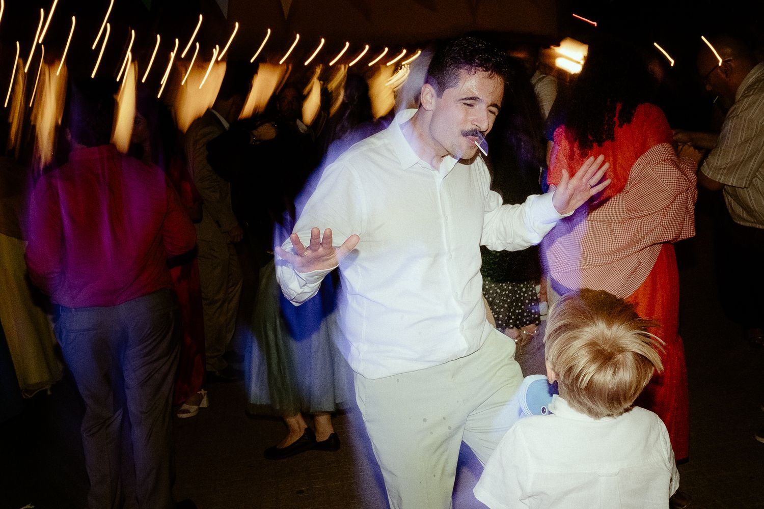 Man with mustache dancing in white shirt at a lively party with colorful lights and guests in background.