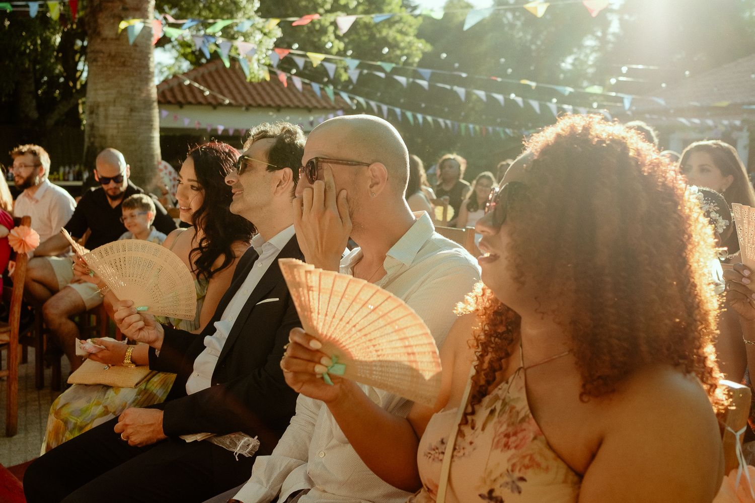 Guests at an outdoor wedding ceremony hold decorative fans while seated, bathed in warm golden sunlight.