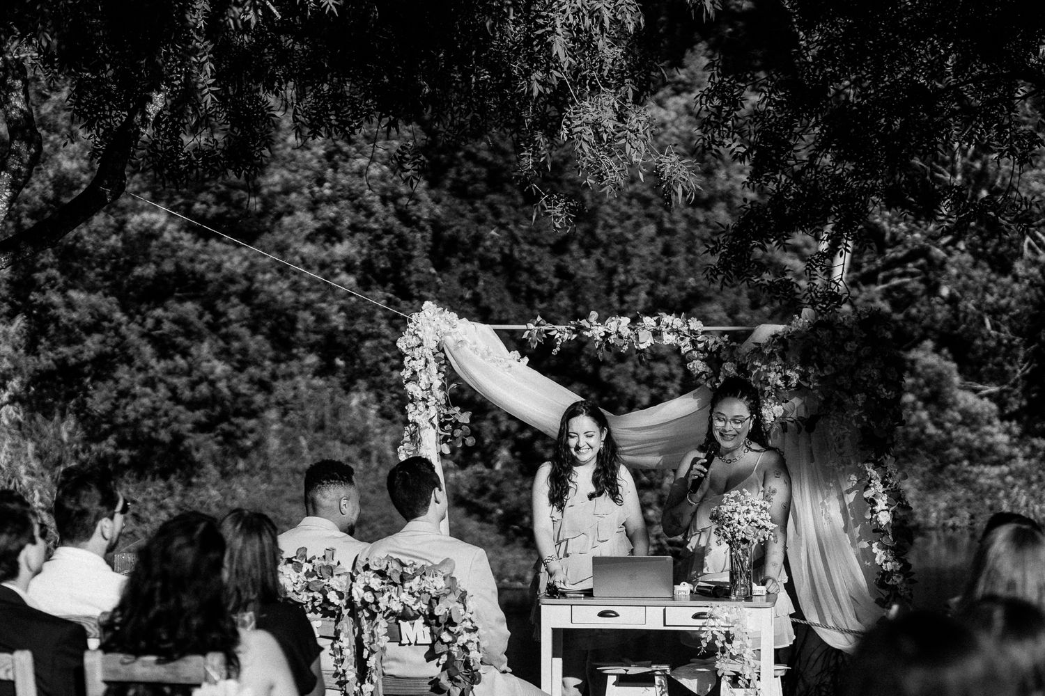Black and white outdoor wedding ceremony with guests seated before a floral arch and two women speaking at a table.
