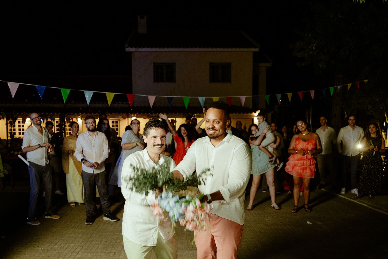 Two grooms walk together at outdoor wedding reception with colorful bunting lights and guests celebrating at night.