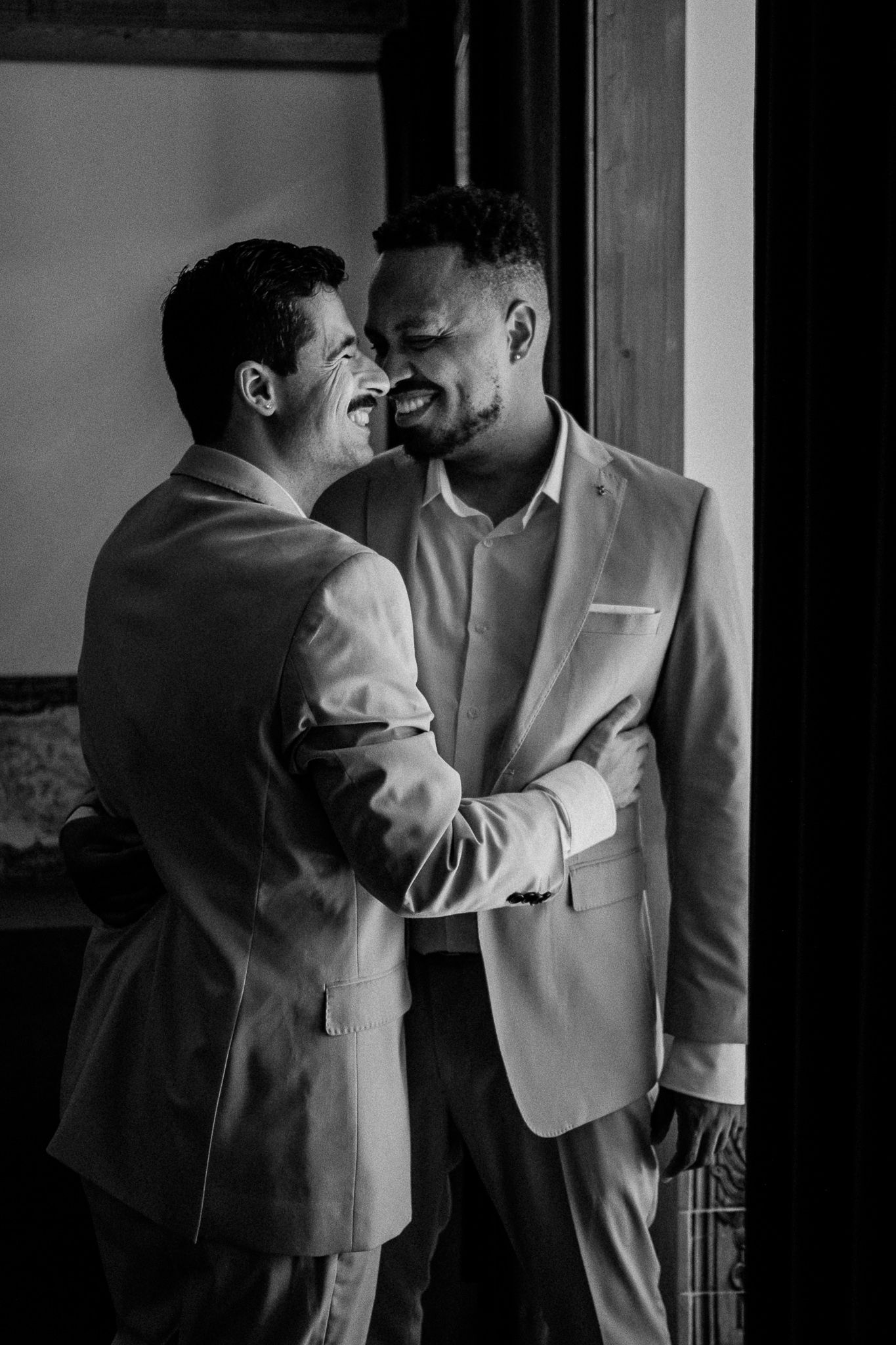 Black and white photo of two grooms in suits sharing a joyful moment, laughing and embracing at their wedding.