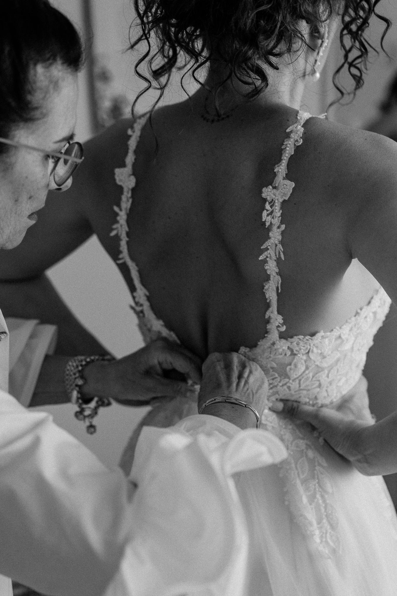 Black and white photo of bride having her lace wedding dress fastened by two helpers during bridal preparations.