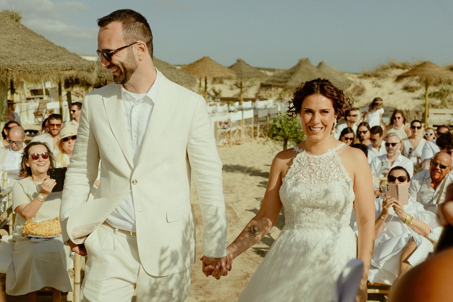 Happy newlywed couple holding hands walking at outdoor beach wedding ceremony with guests applauding behind them.