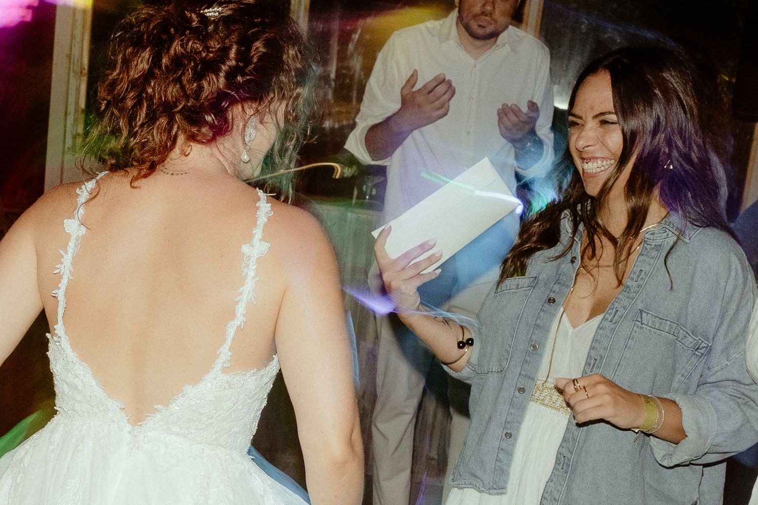 Bride in white lace gown laughing with guest in denim jacket at colorful wedding reception.