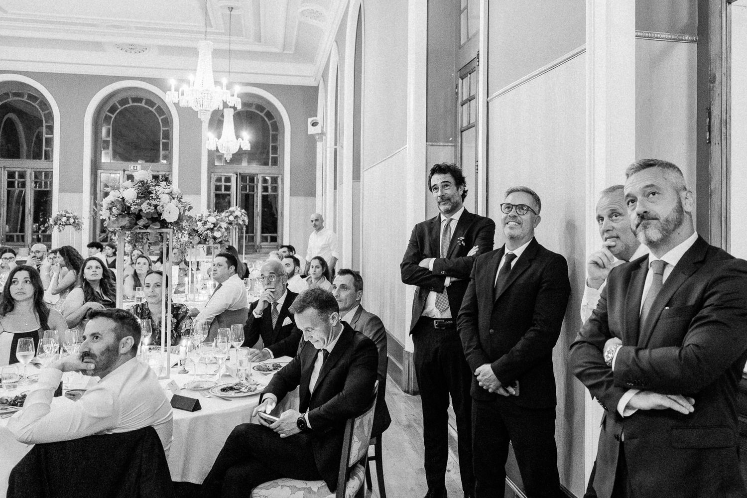 Black and white wedding reception photo showing elegantly dressed guests standing and seated in a grand ballroom.