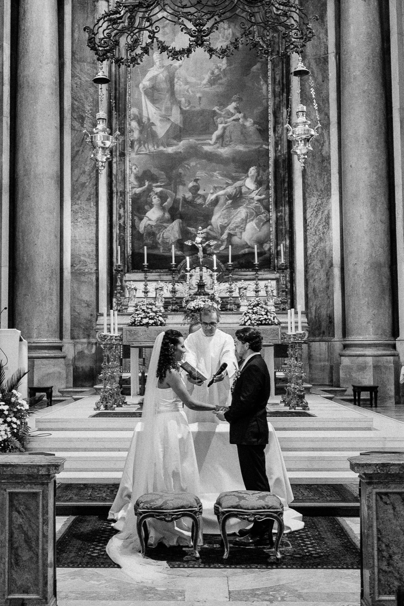 Black and white photo of a bride and groom exchanging vows at a grand church altar with ornate decor.