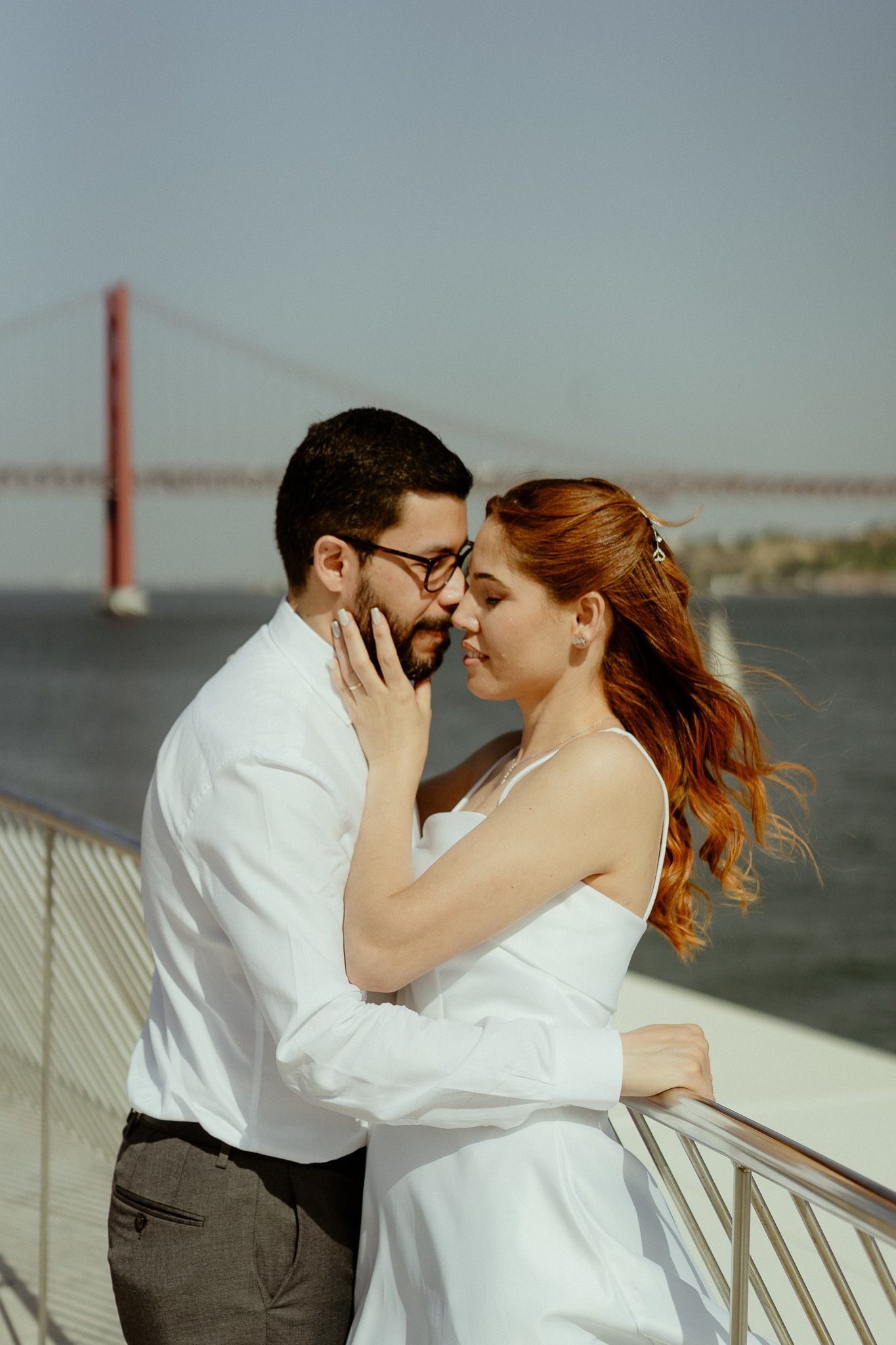 Couple in wedding attire embracing near waterfront railing with red suspension bridge in background.