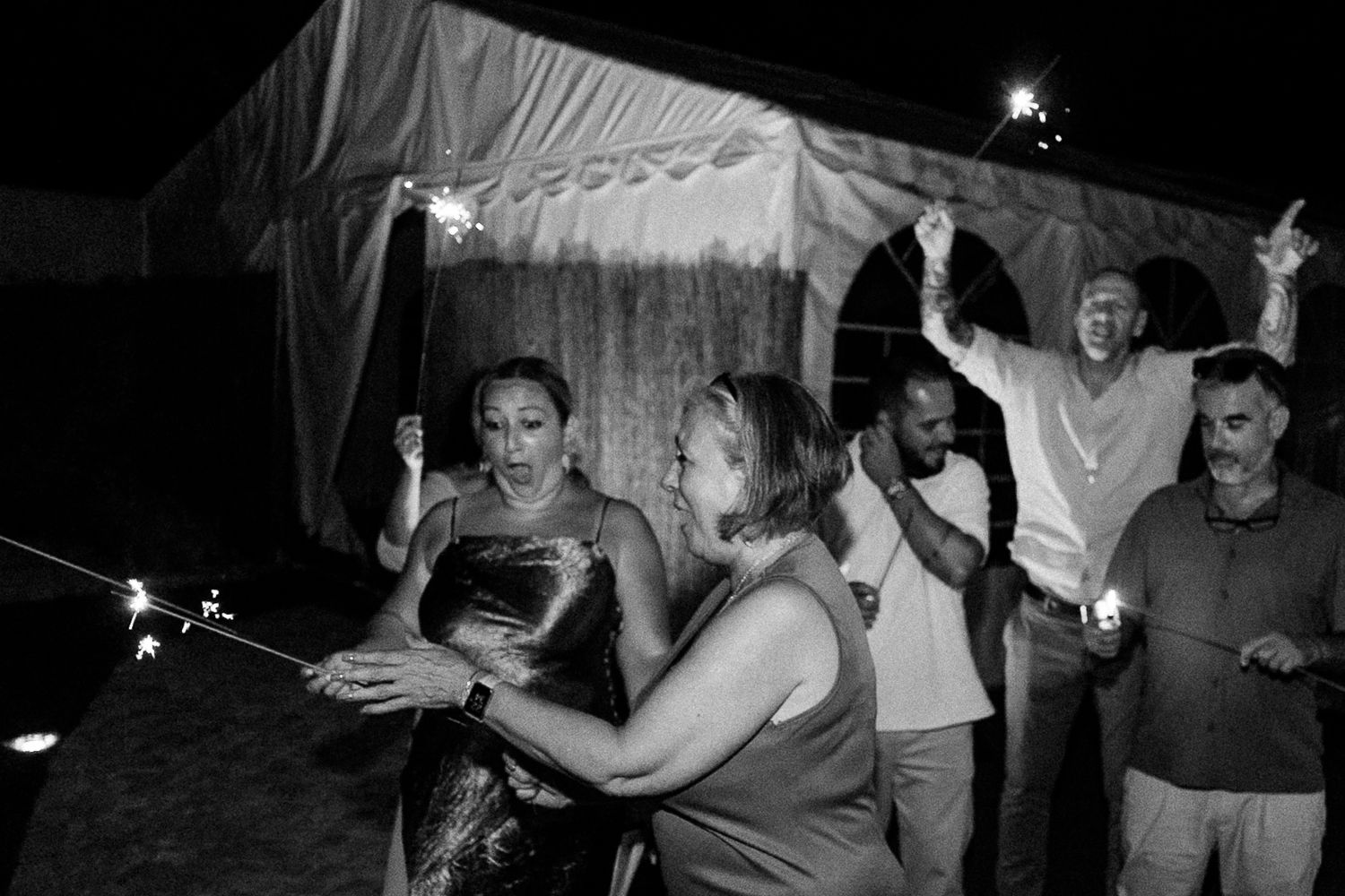 Black and white photo of guests celebrating outdoors at night with sparklers near a tent.
