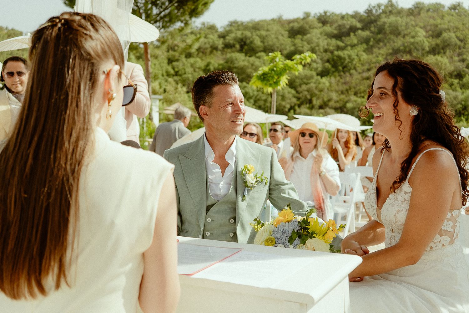 Bride and groom smiling at outdoor wedding ceremony with guests in background on sunny day.