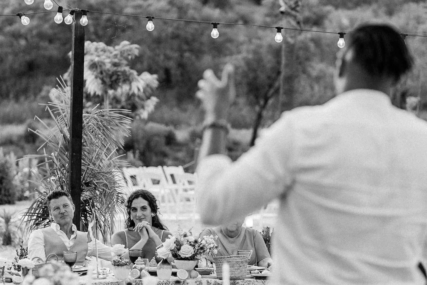 Black and white photo of wedding guests listening to a toast at an outdoor reception with string lights.
