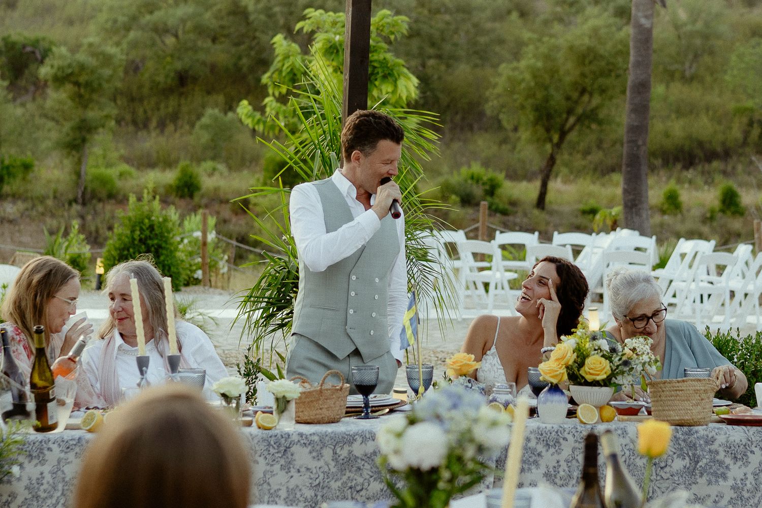 Man giving a toast speech at an outdoor wedding reception dinner table with guests smiling.
