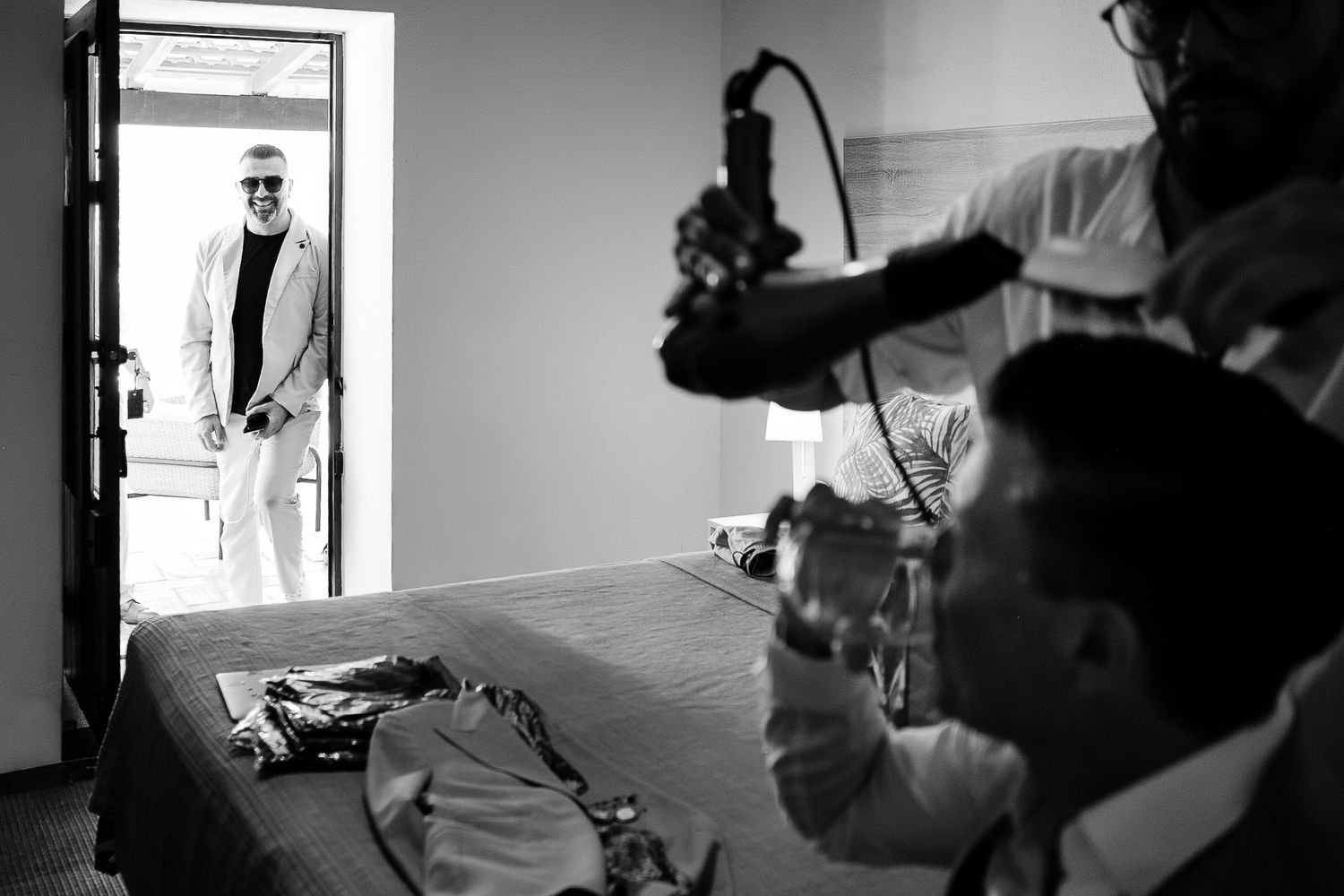 Black and white behind-the-scenes photo of a film crew filming a stylish man in sunglasses standing in a doorway.
