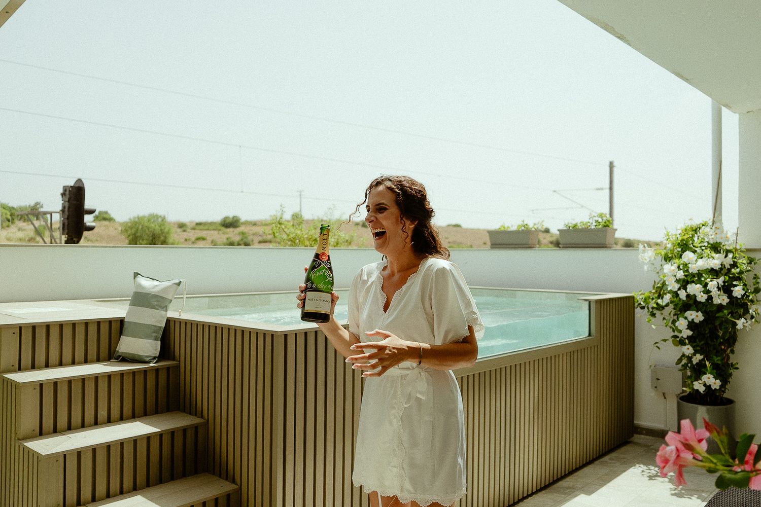 Woman in white dress holding champagne bottle by outdoor hot tub on sunny terrace with ocean view.