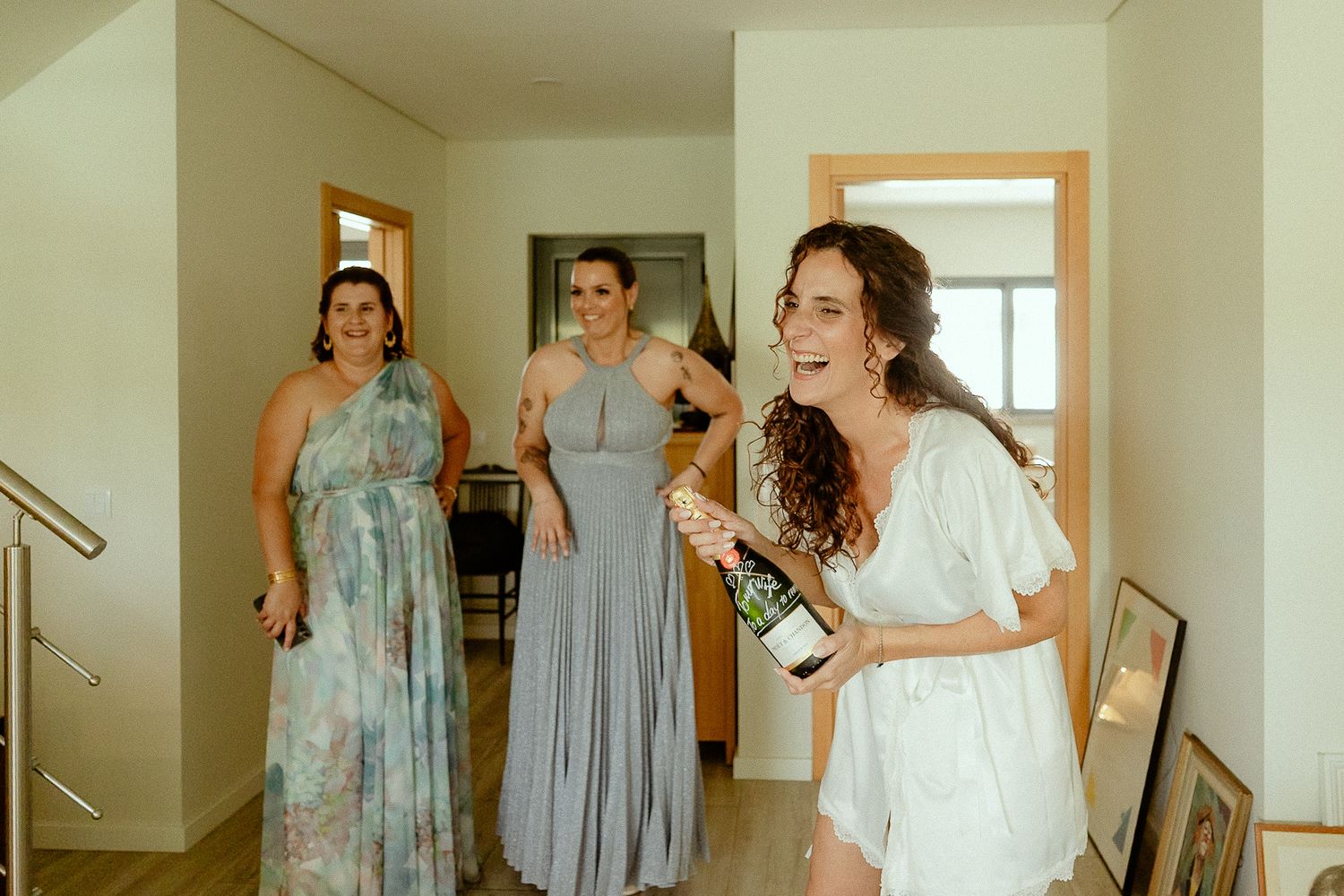 Bride laughing holding champagne bottle with bridesmaids in floral and gray dresses in bright hallway.