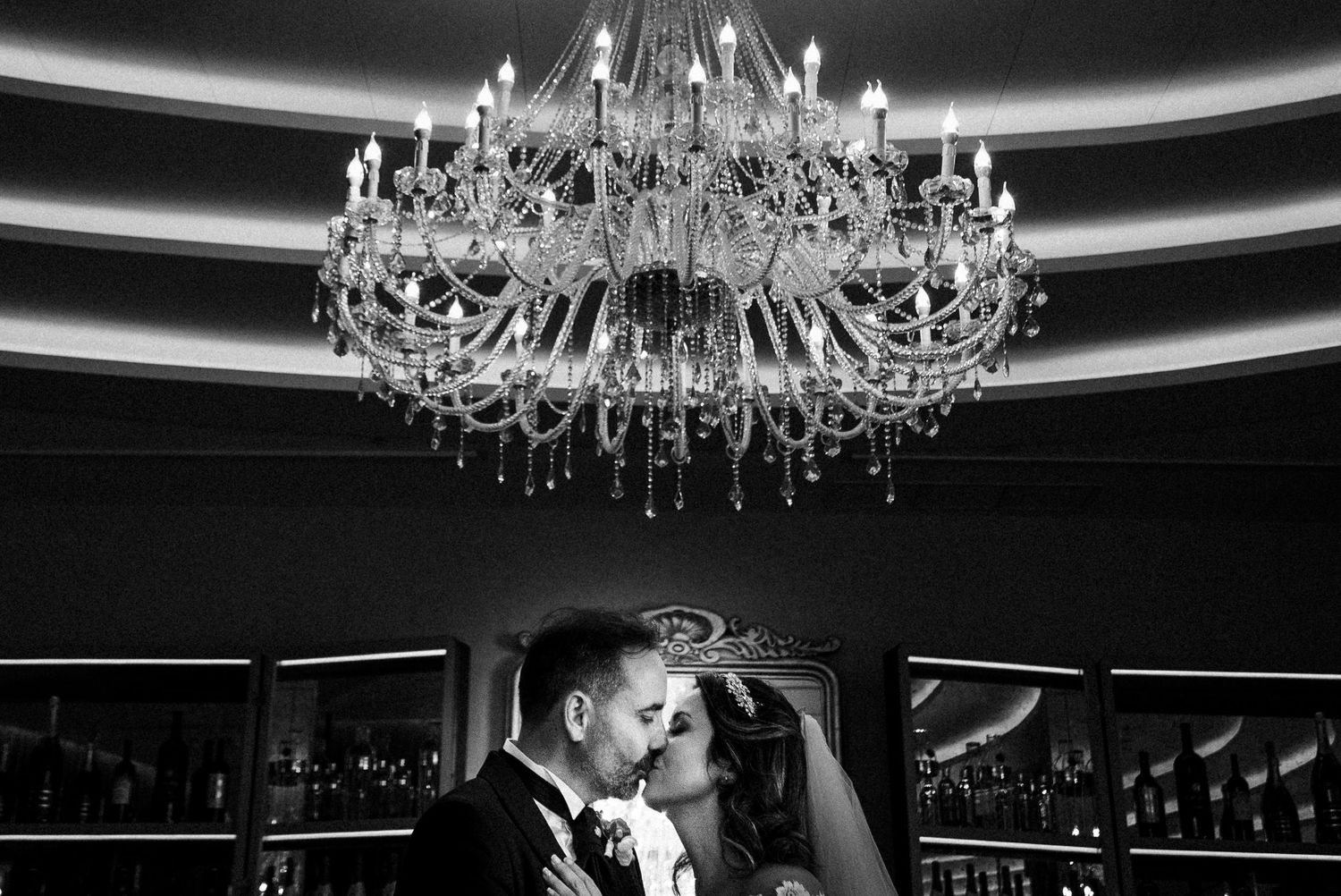 Romantic couple embraces beneath an elegant crystal chandelier in dramatic black and white wedding photography.