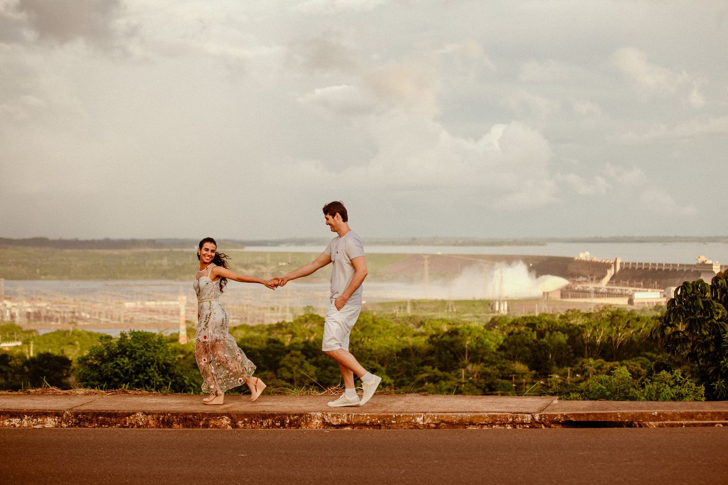 A couple holds hands while balancing and walking along a stone wall overlooking a scenic coastal landscape at sunset.