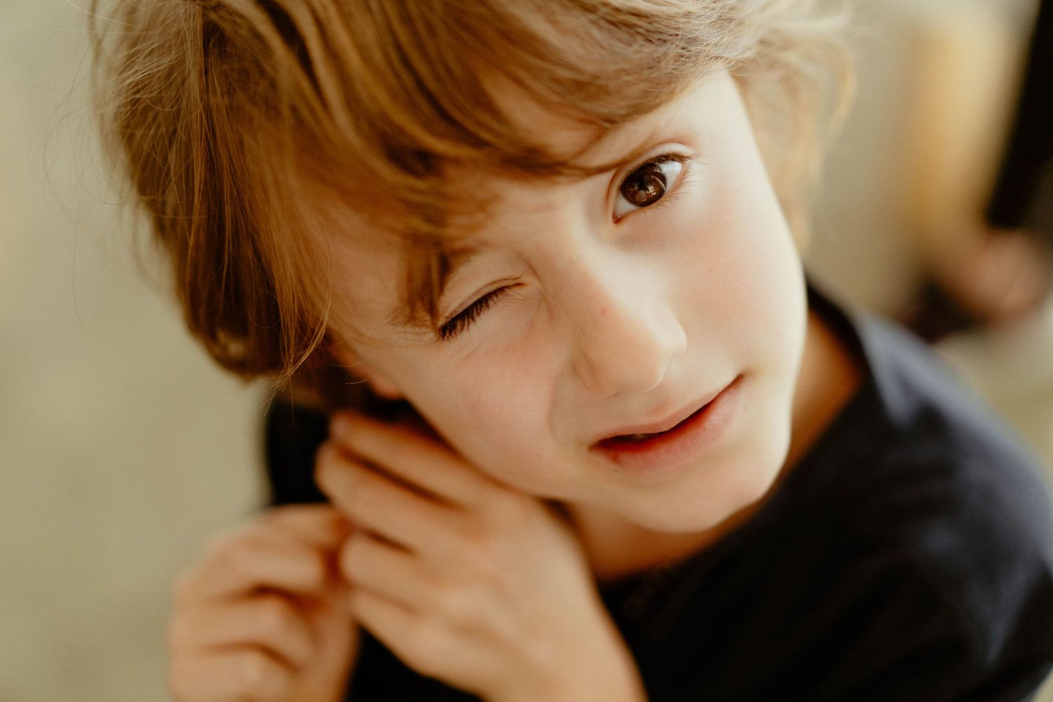 A person with light brown hair wearing a black top winks playfully while touching their face in a warm-lit setting.