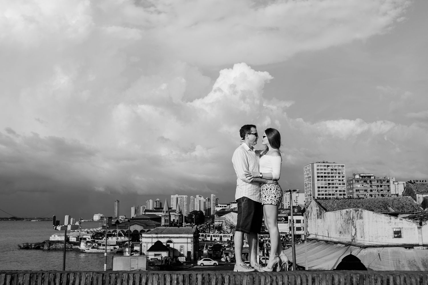 A romantic couple embraces on a rooftop overlooking a cityscape with dramatic clouds in black and white.
