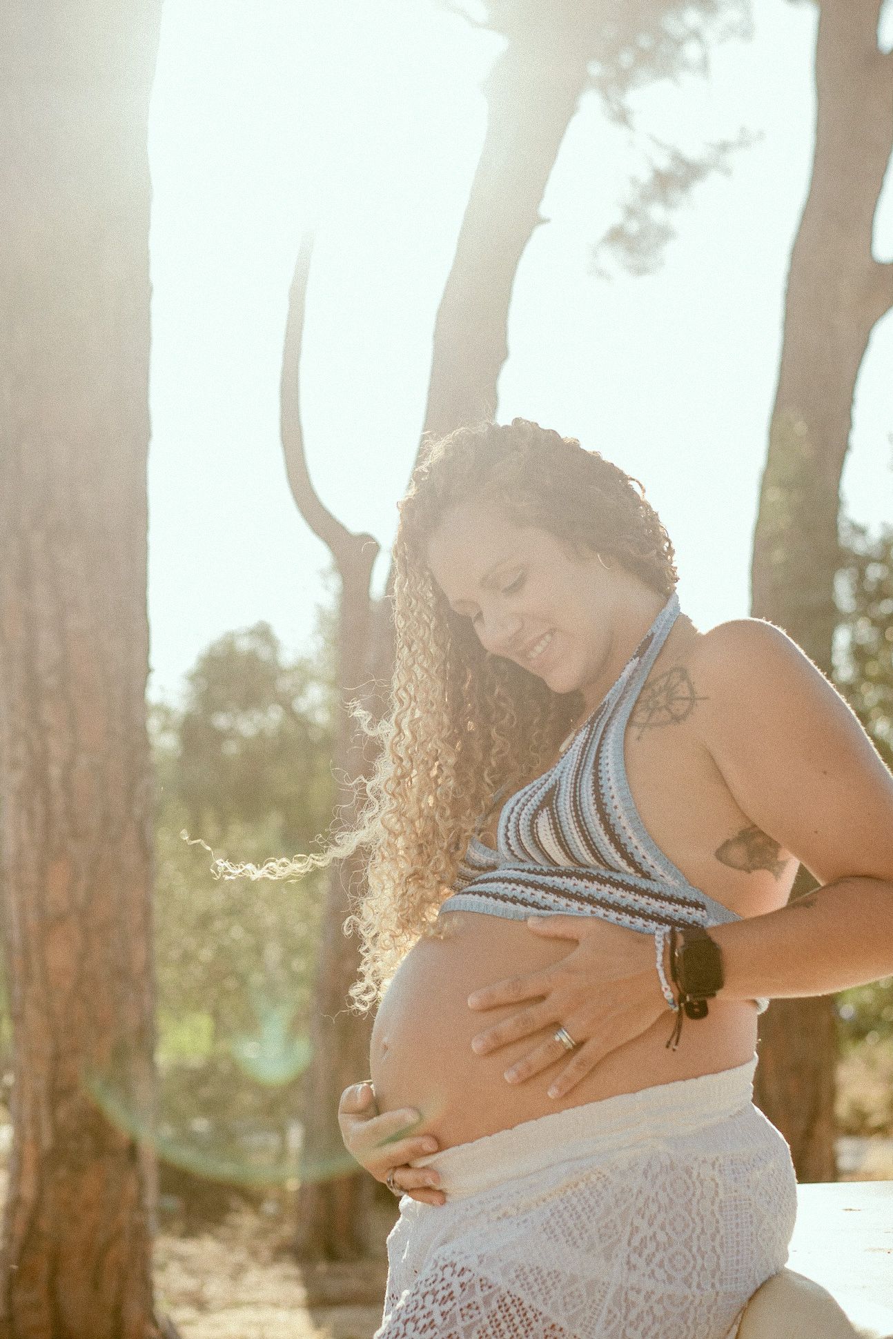 A pregnant woman in a striped top caresses her belly in a sunlit forest setting.