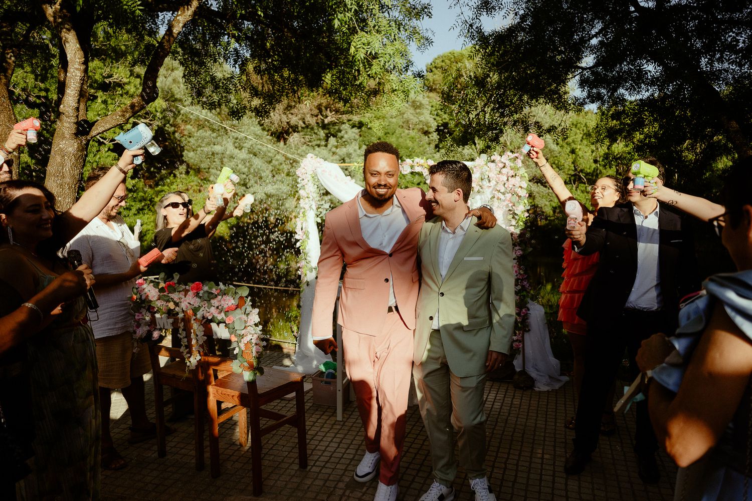 Two grooms in pink and green suits celebrate their wedding exit through cheering guests outdoors.