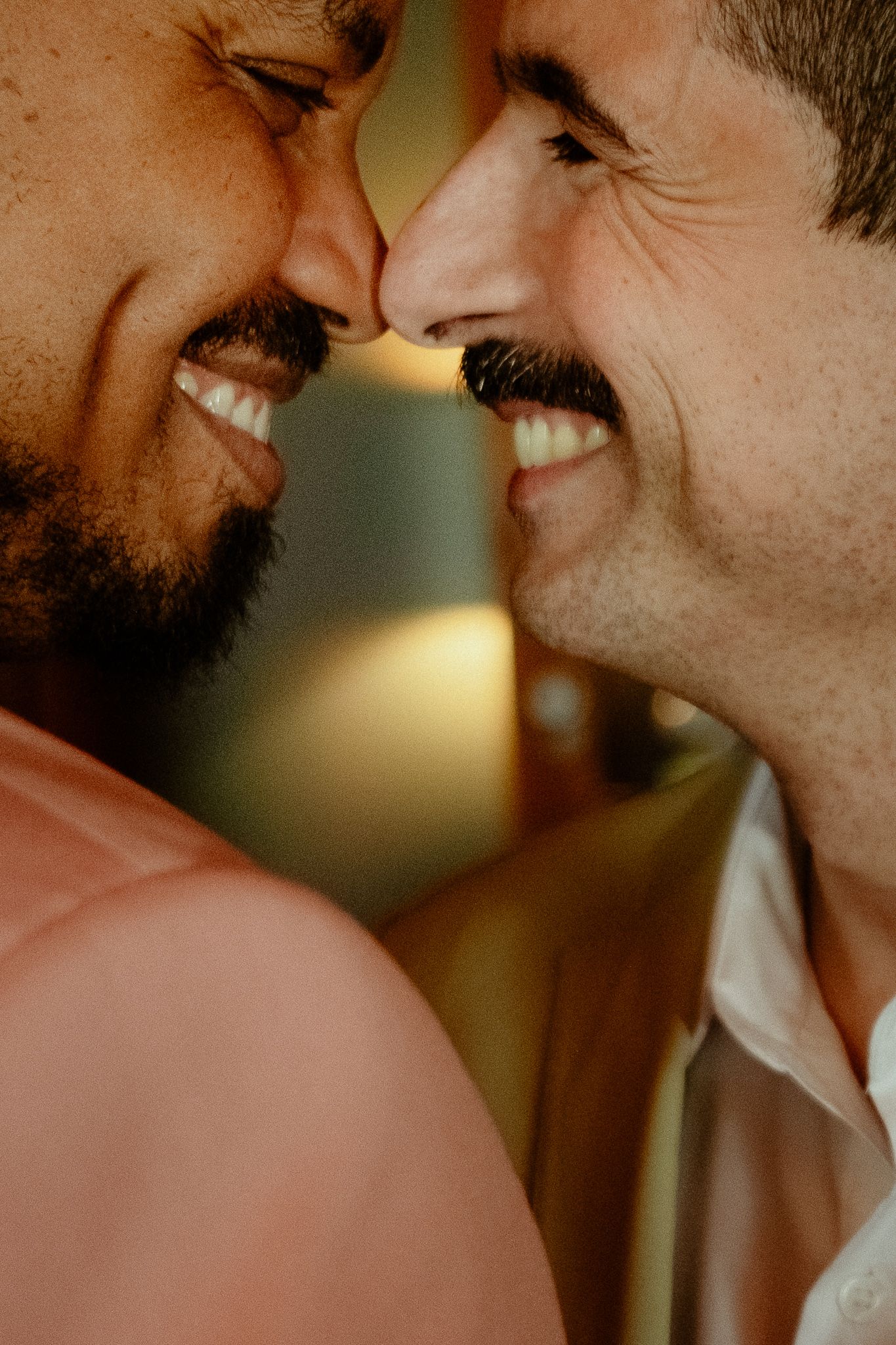 Two smiling men facing each other closely, sharing an intimate moment, one with a beard and one with a mustache.