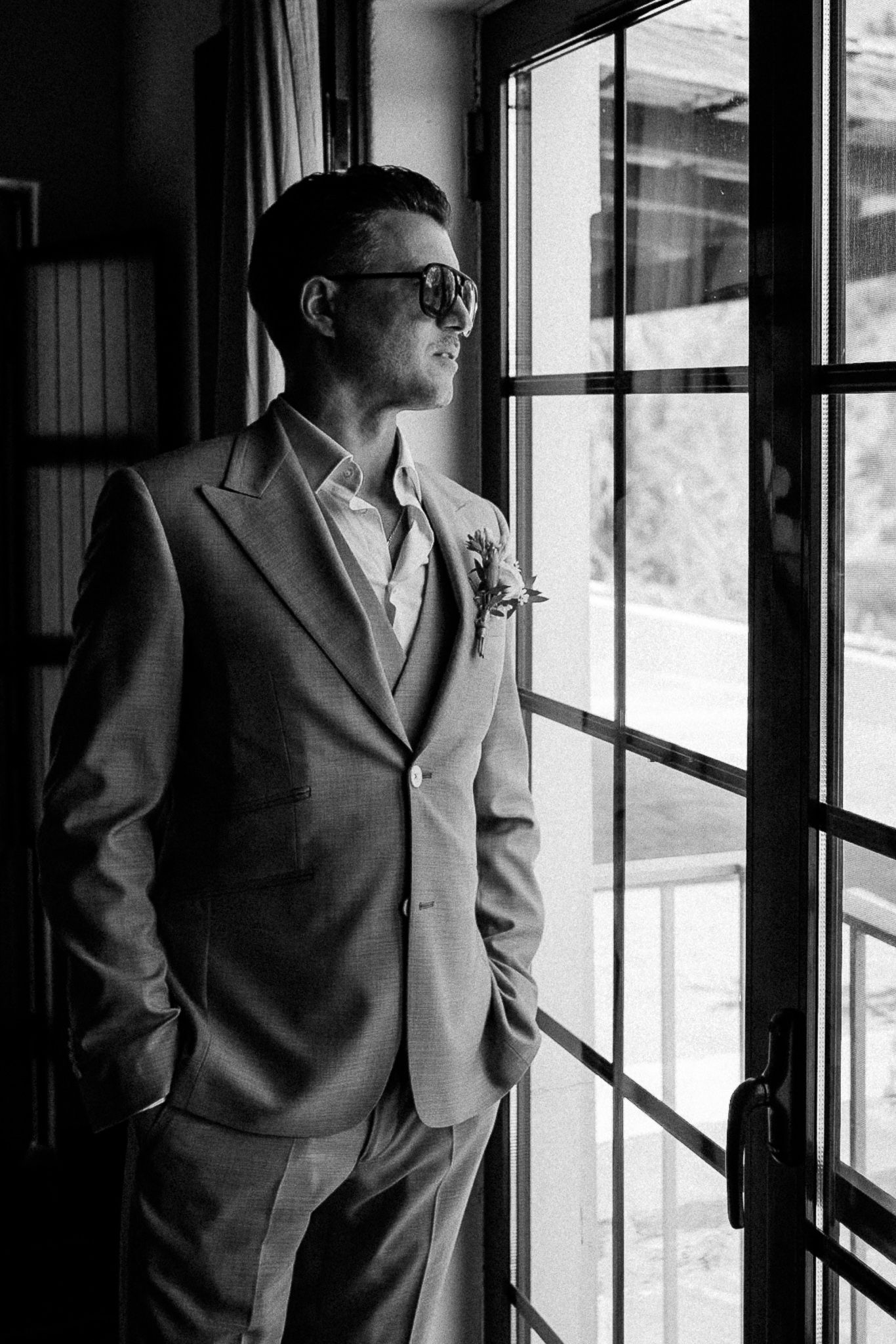 Elegant groom in gray suit and sunglasses standing by a window with hands in pockets.