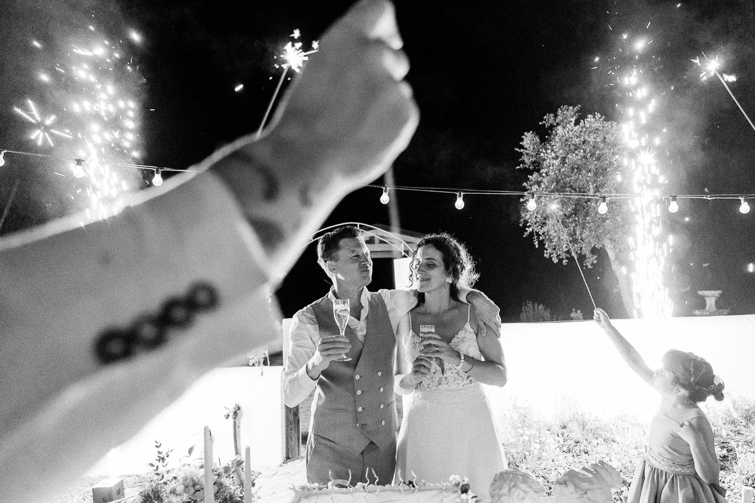 Black and white wedding photo of couple celebrating with sparklers at night outdoor reception.