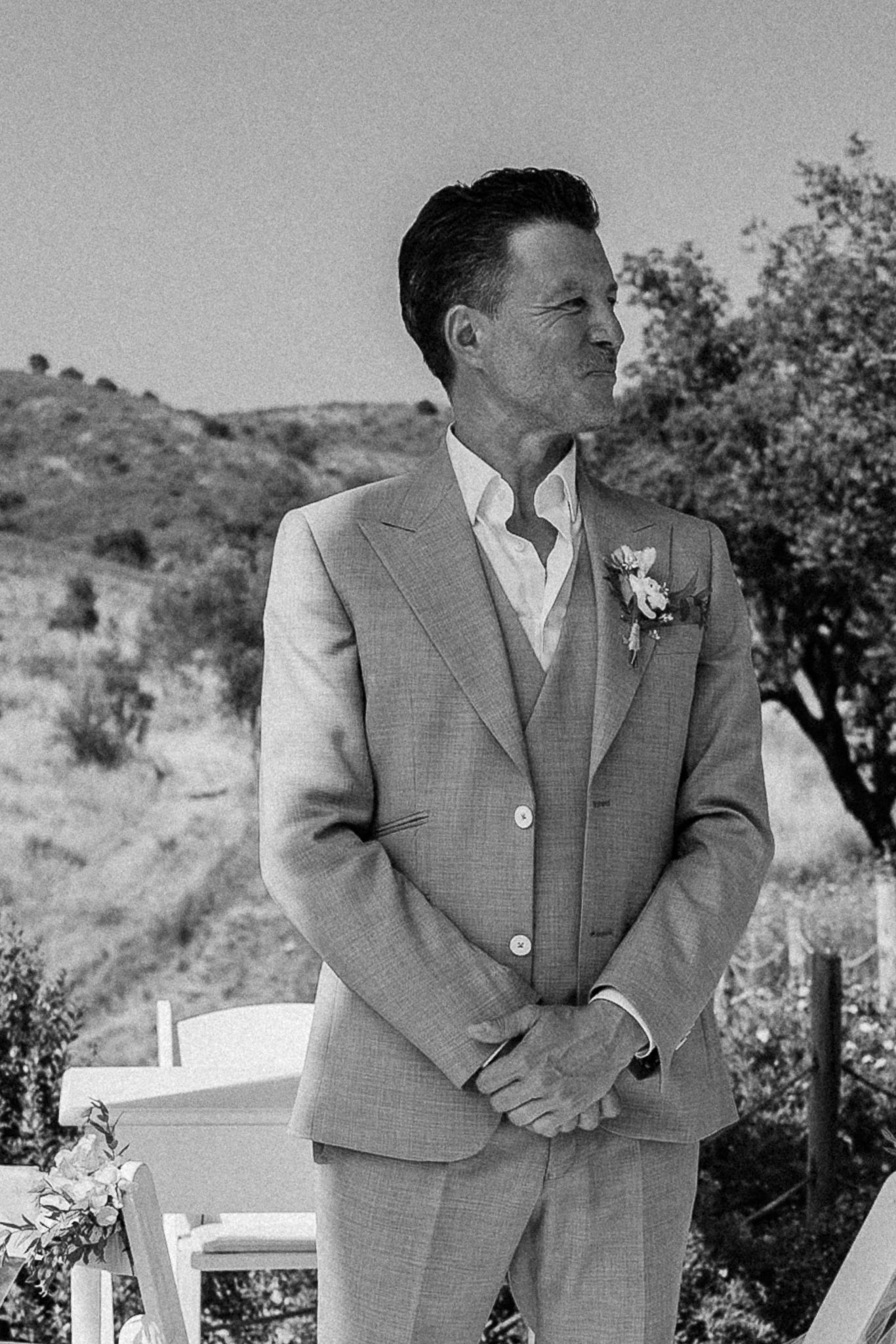 A groom in a light suit with boutonniere stands outdoors at a wedding ceremony with hills in the background.