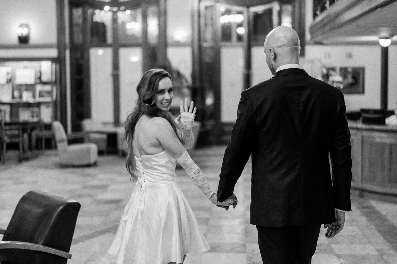 Black and white photo of a bride in a white dress waving while holding hands with a groom in a black suit.