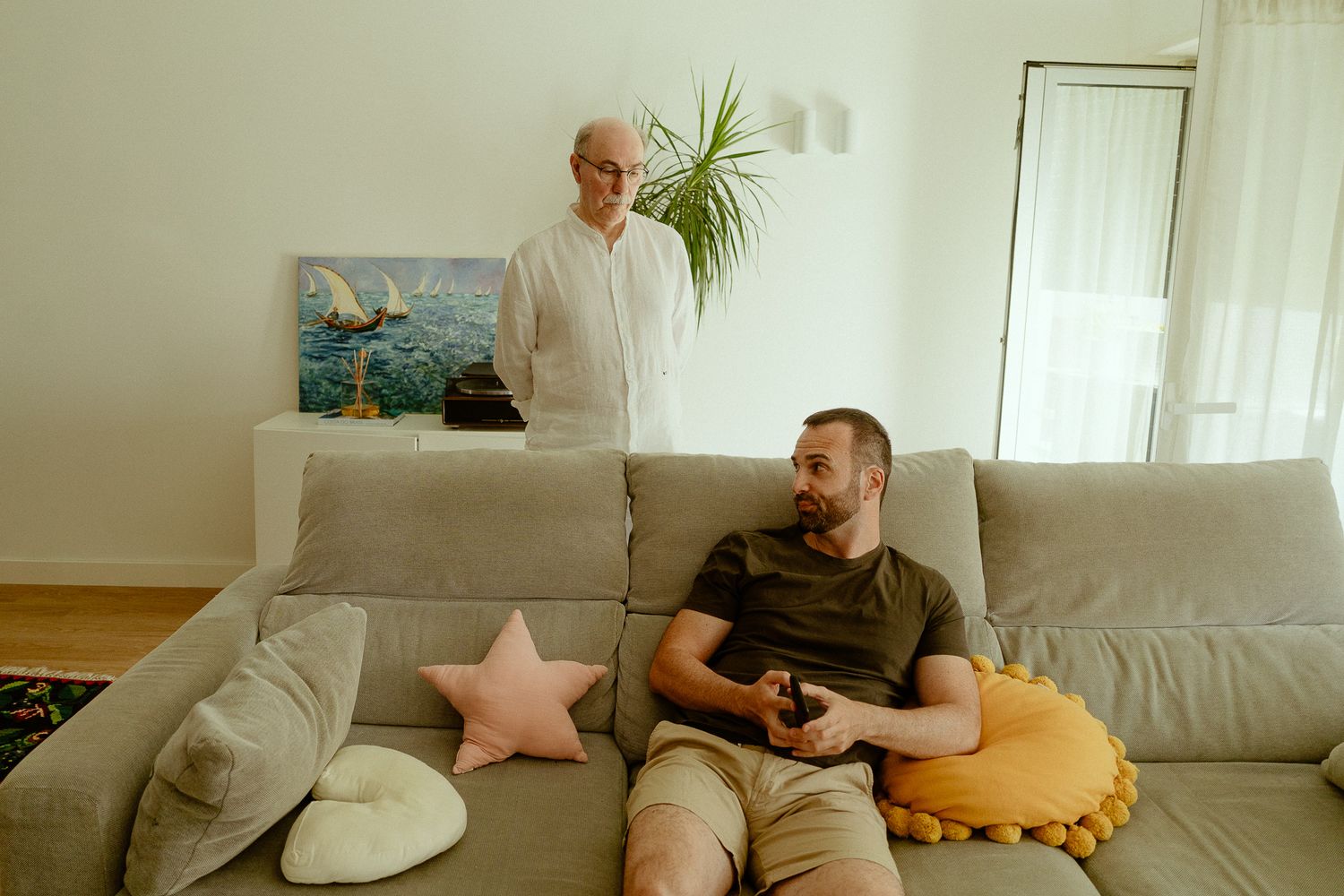 Two men relaxing in a modern living room, one seated on a gray sofa holding a remote, another standing nearby.