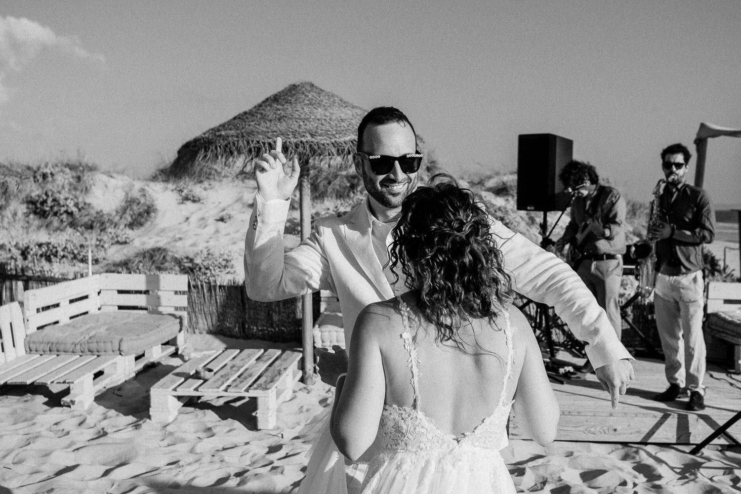 Bride and groom share first dance on sandy beach at outdoor wedding reception with live band performing in background.