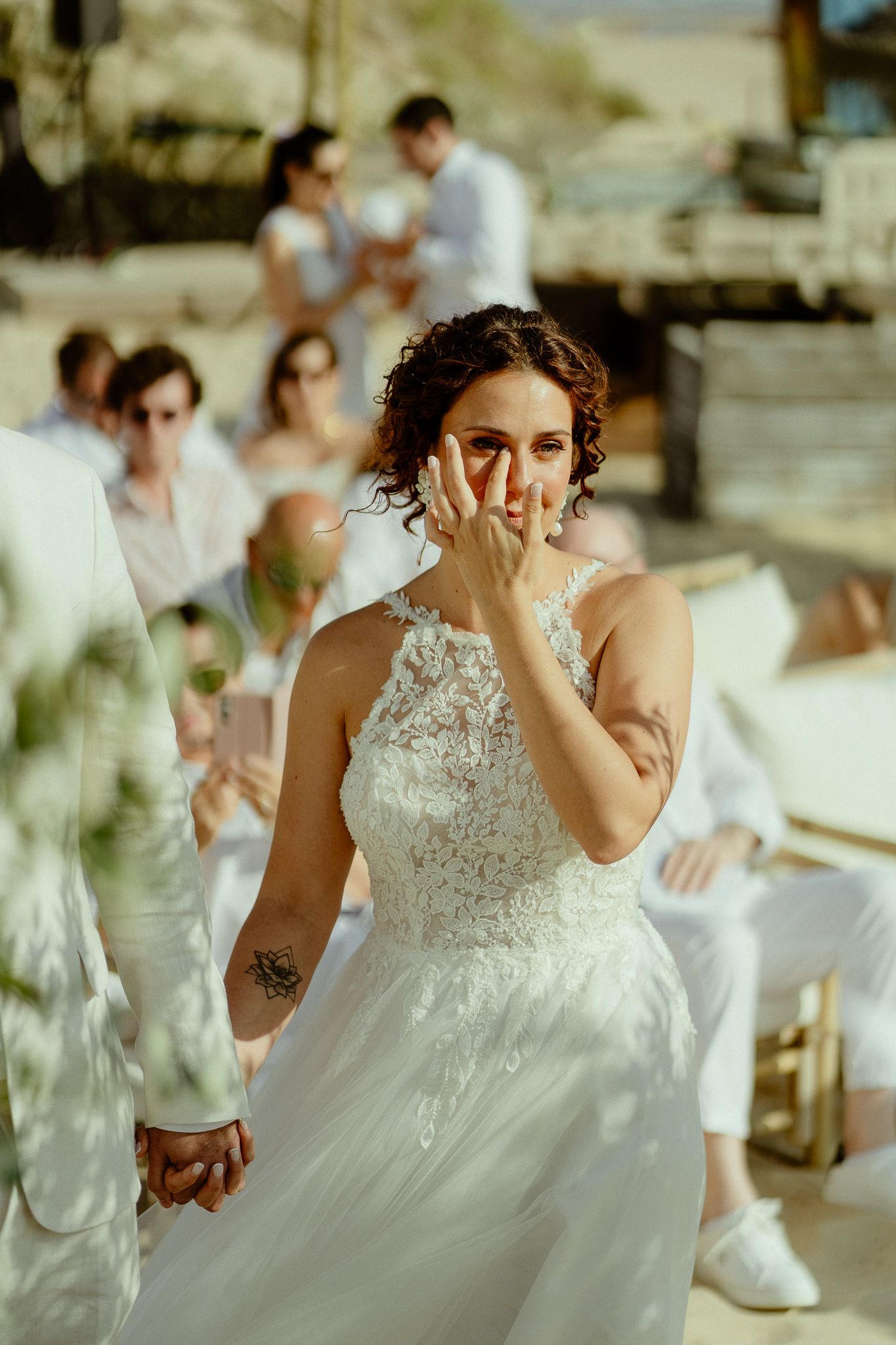 Emotional bride in lace wedding dress wiping tears during outdoor ceremony with guests seated in background.
