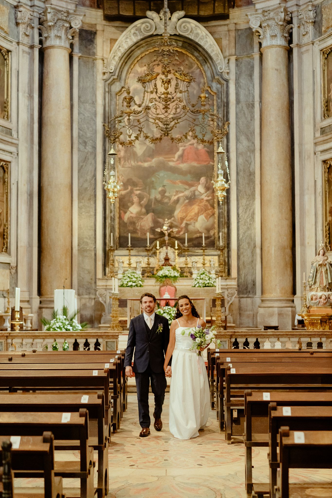 Bride and groom walking down the aisle of an ornate Baroque church with a grand altar painting.