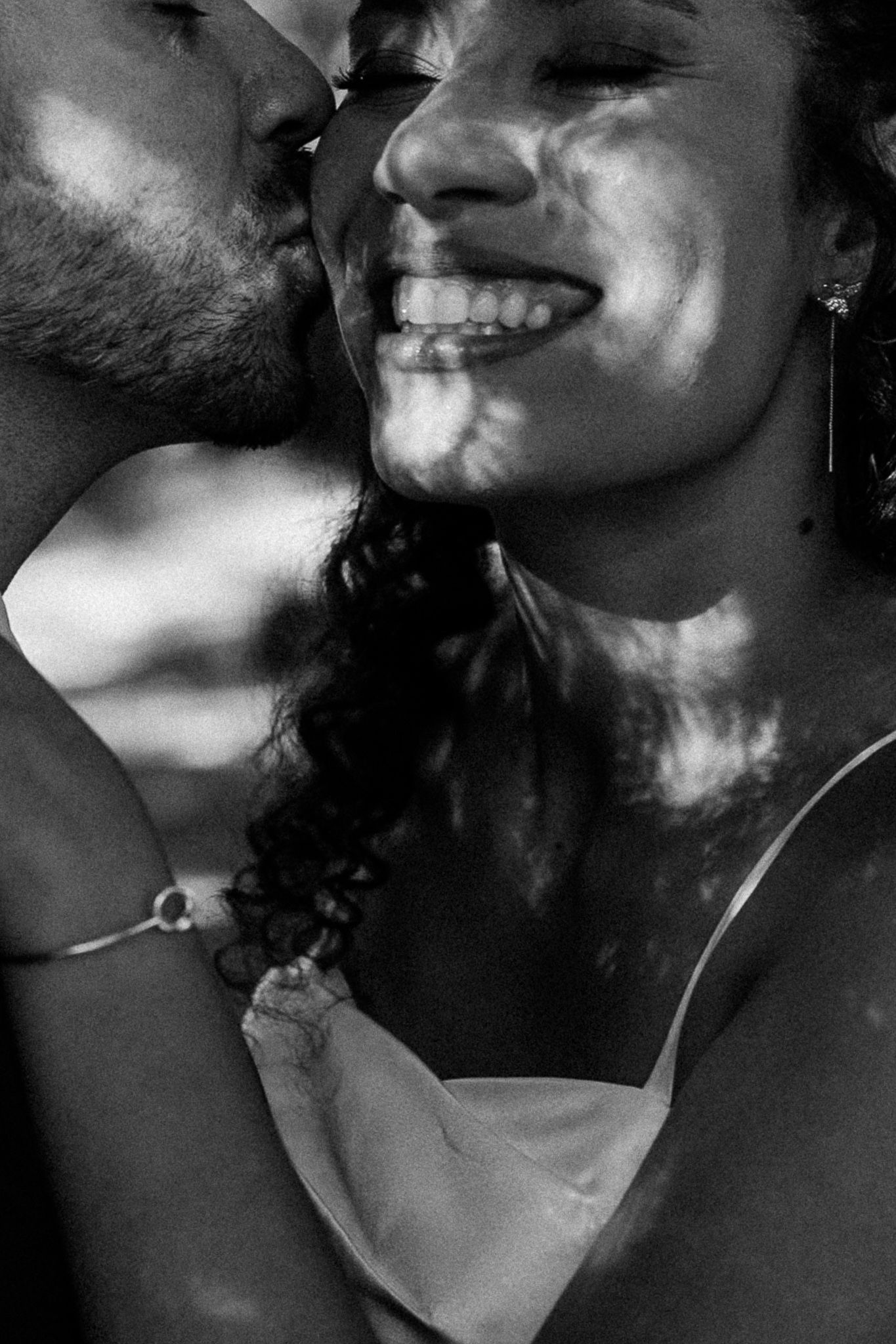 Black and white photo of a couple sharing a tender moment, man kissing smiling woman's cheek in dappled light.
