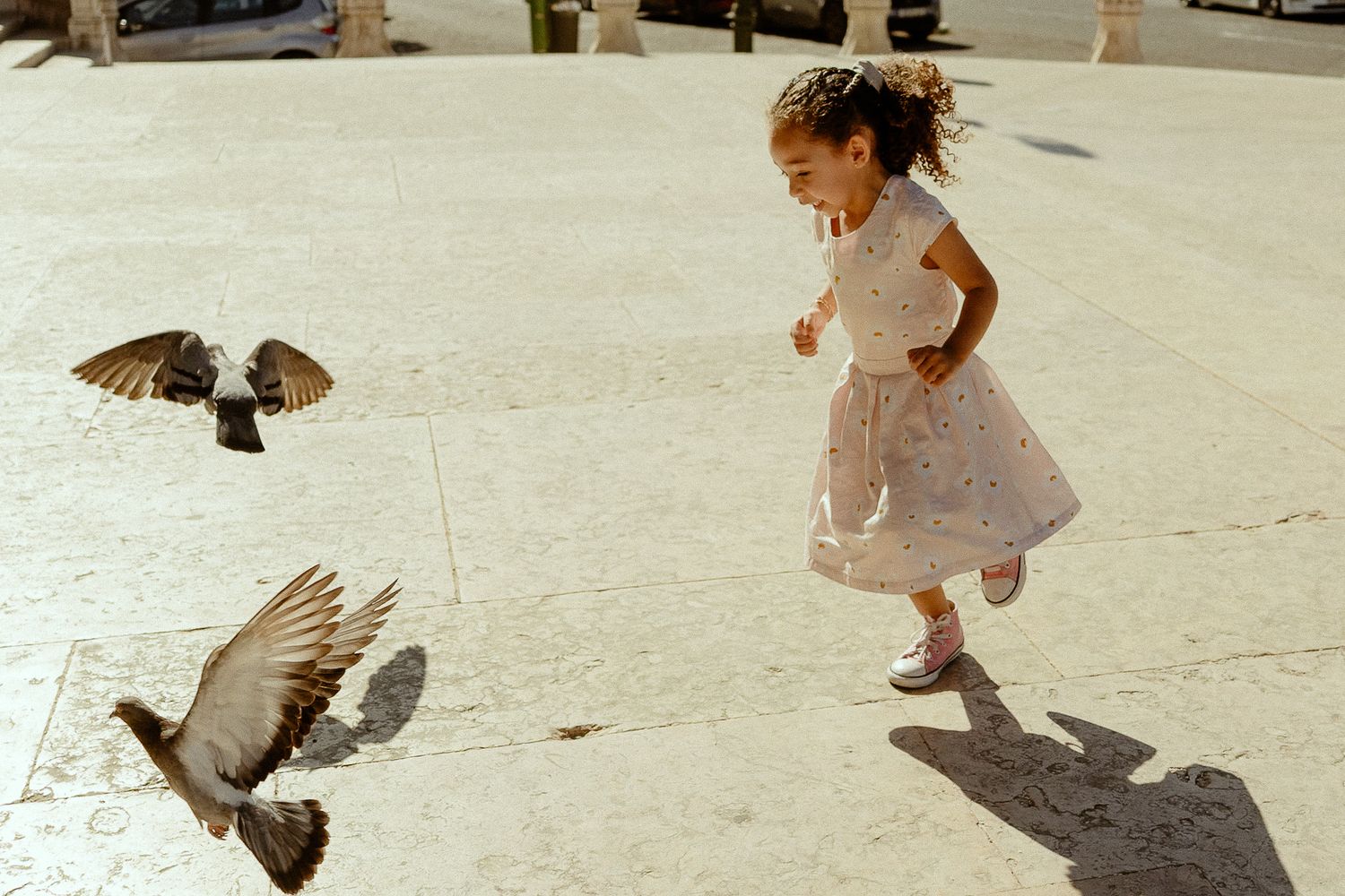 A young girl in a white dress chases pigeons taking flight on a sunlit plaza.