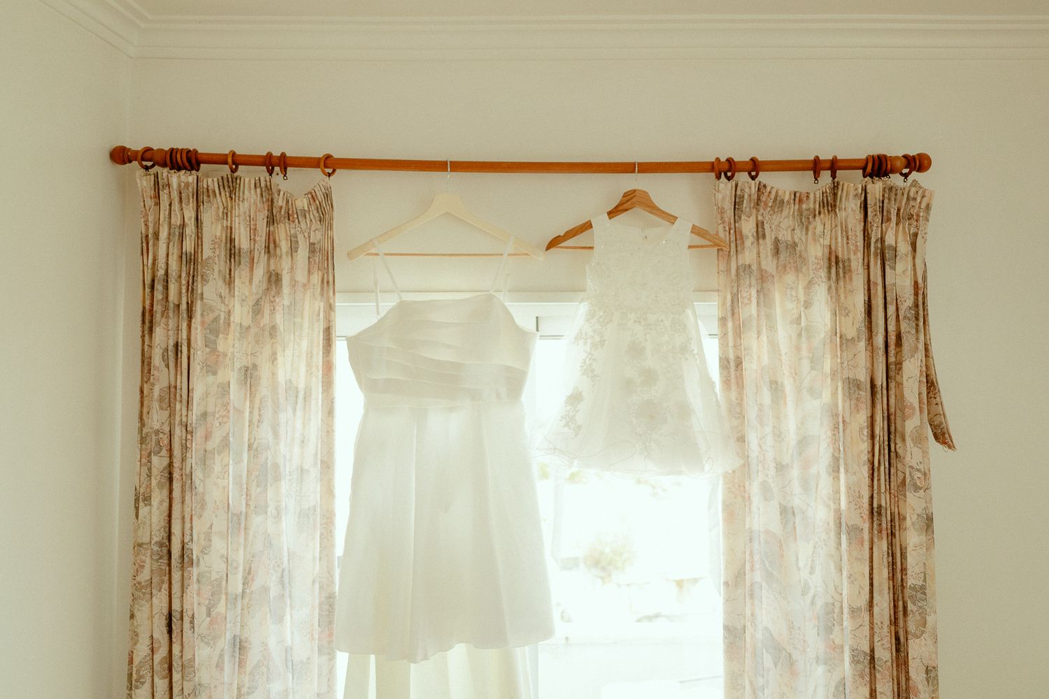 Two white dresses hanging on a curtain rod in front of a sunlit window with floral curtains.