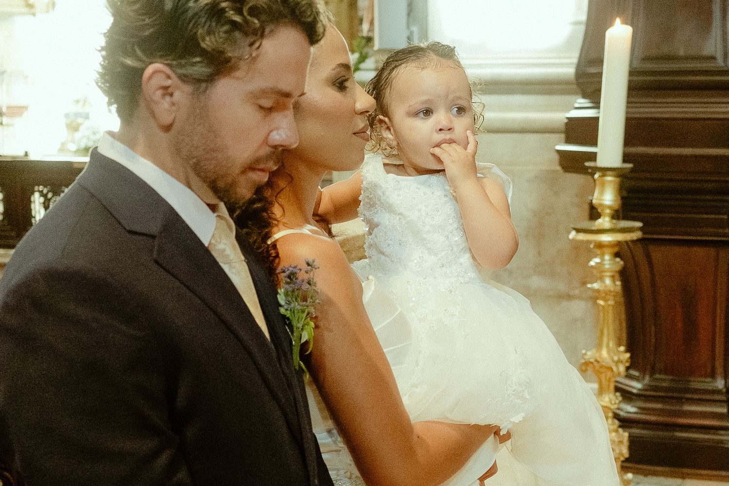 A couple in wedding attire shares a tender moment with their young daughter in a white dress inside a church.