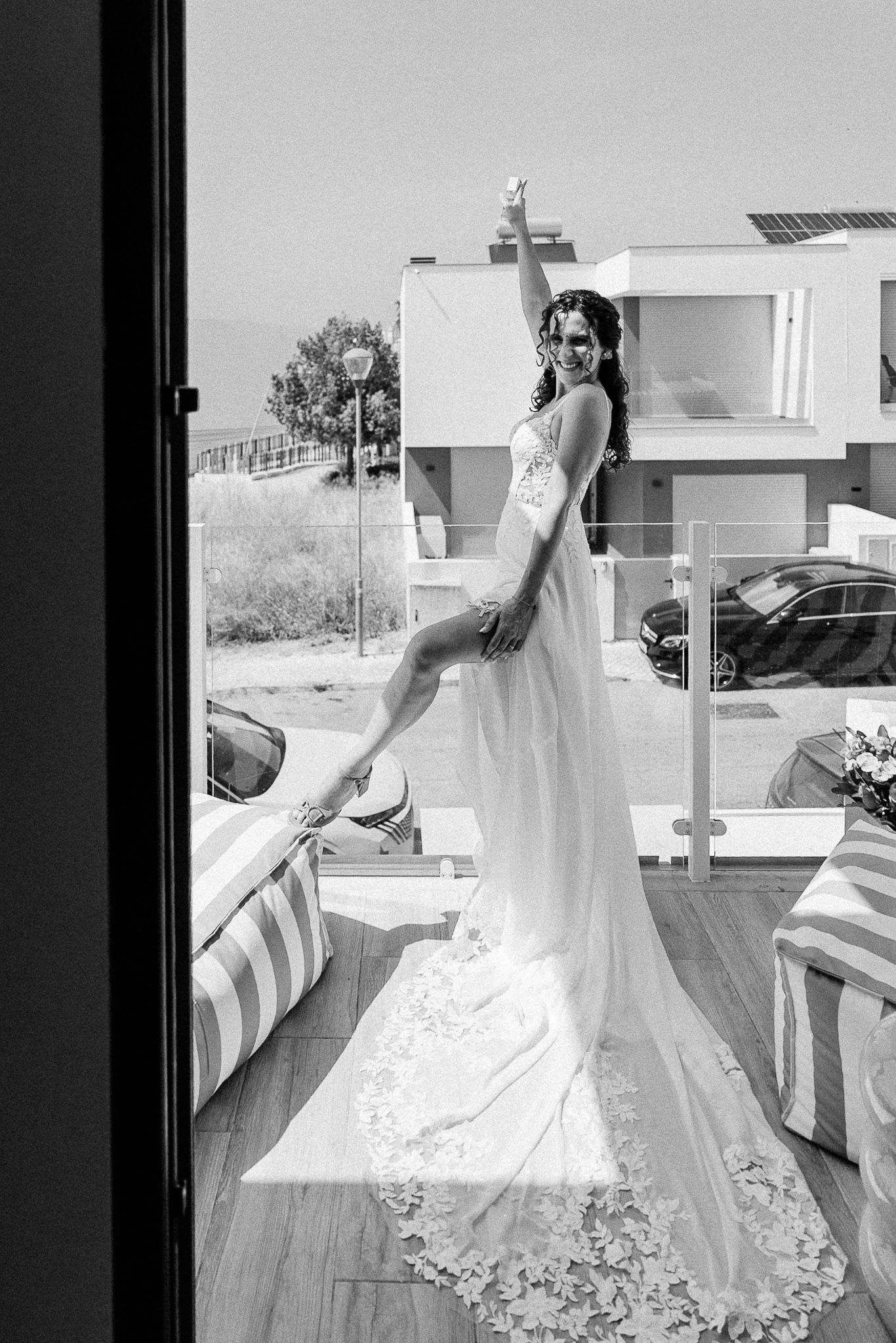 Bride in lace wedding gown poses gracefully on a sunlit balcony, one arm raised, with a floral train.